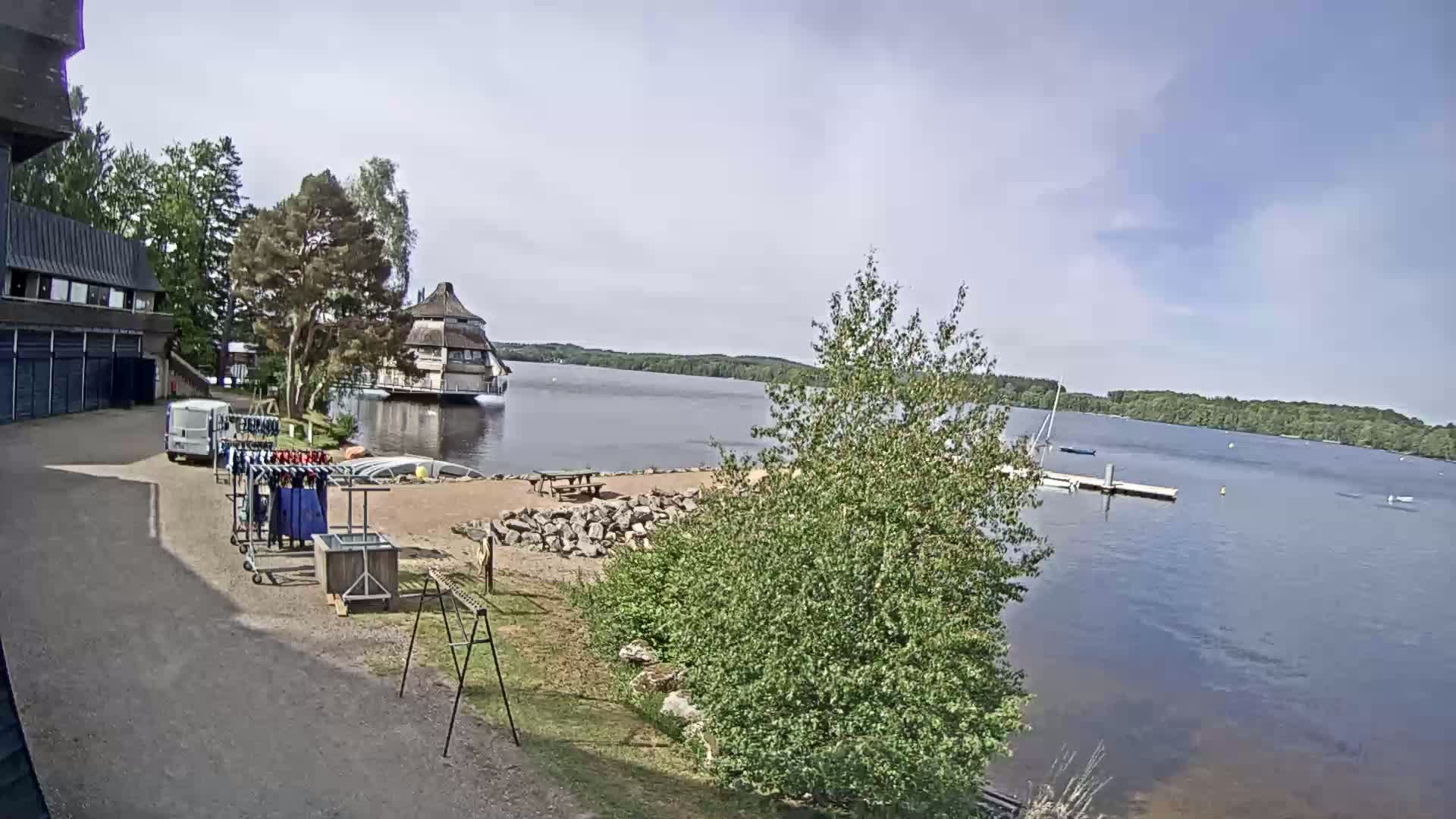 A lakeside view on a partly cloudy day shows a building, a small sandy beach area with a picnic table, and wetsuits drying on racks near a van.