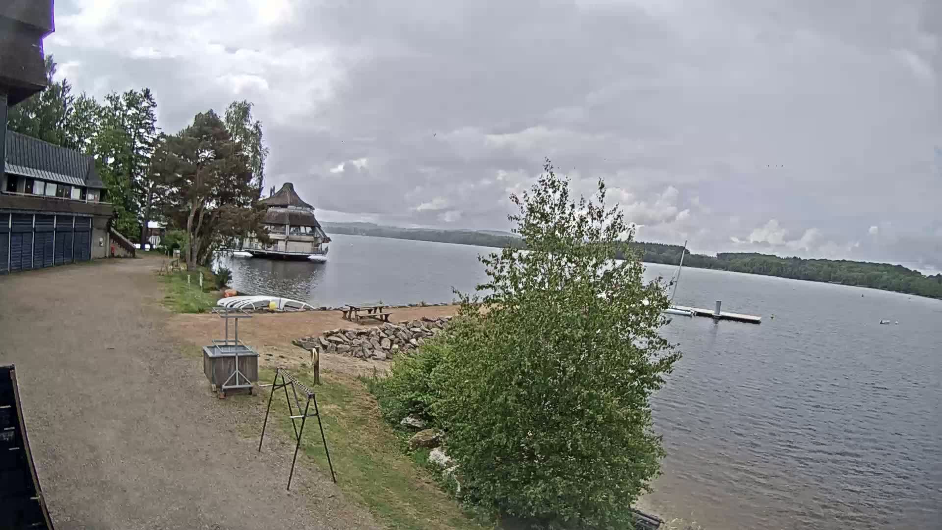 A lakeside view on an overcast day shows a building with a peaked roof over the water, a gravel area with picnic tables and a small boat dock, and lush green vegetation.