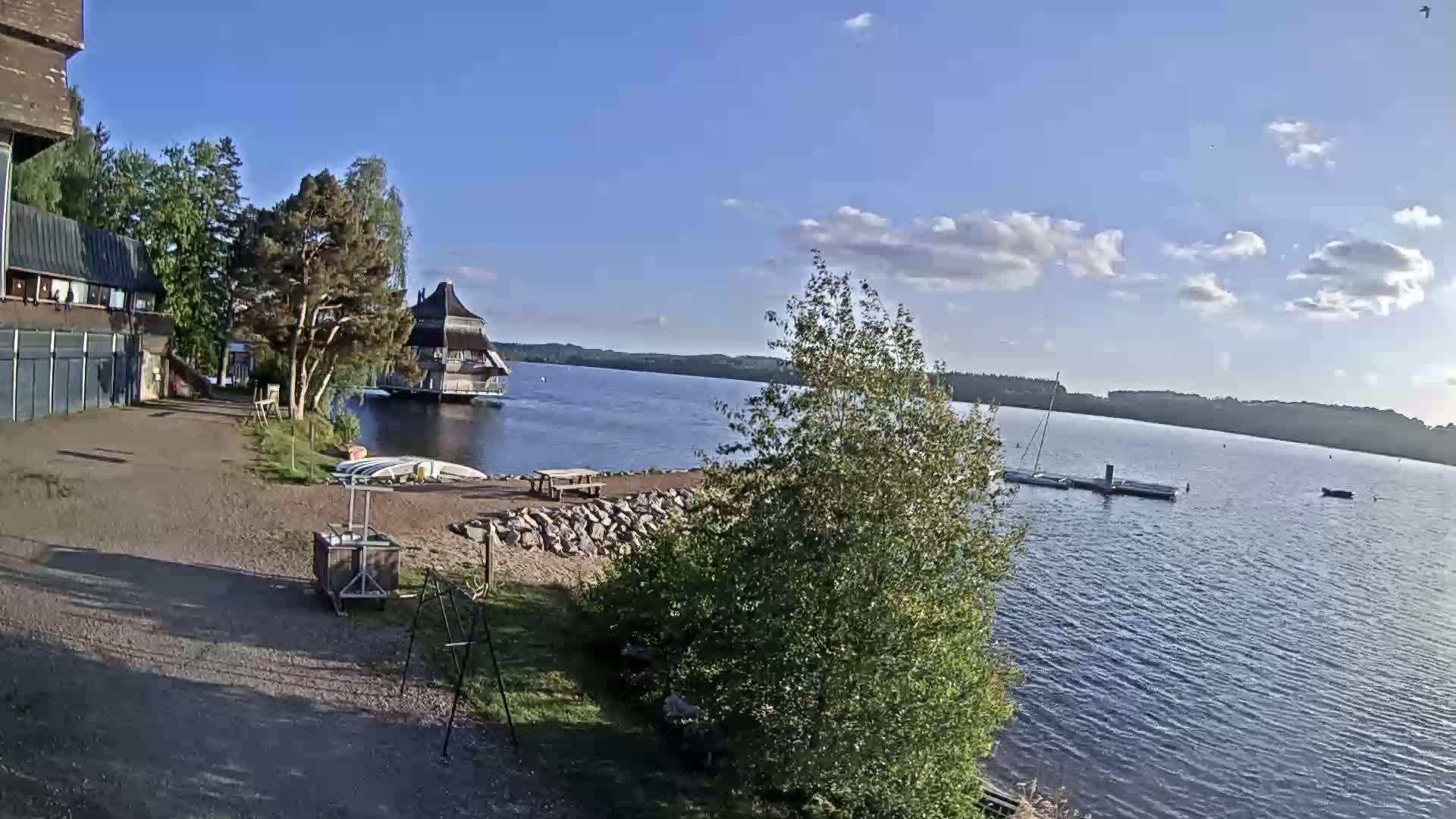 A calm lake is seen under a mostly sunny sky, with a lakeside building, small boats, and a grassy area with picnic tables.