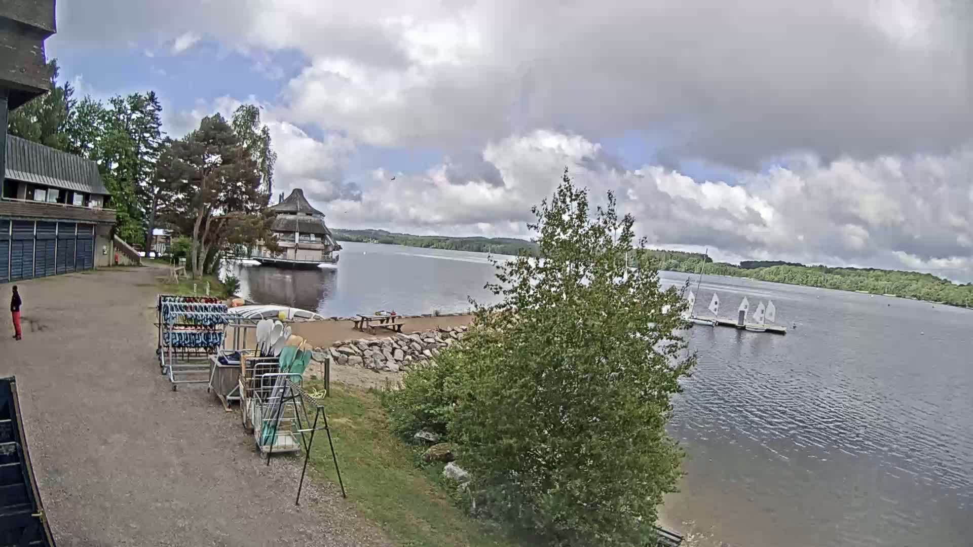 A lakeside view on a partly cloudy day shows a building, a dock with sailboats, and various water sports equipment stored near a sandy area.