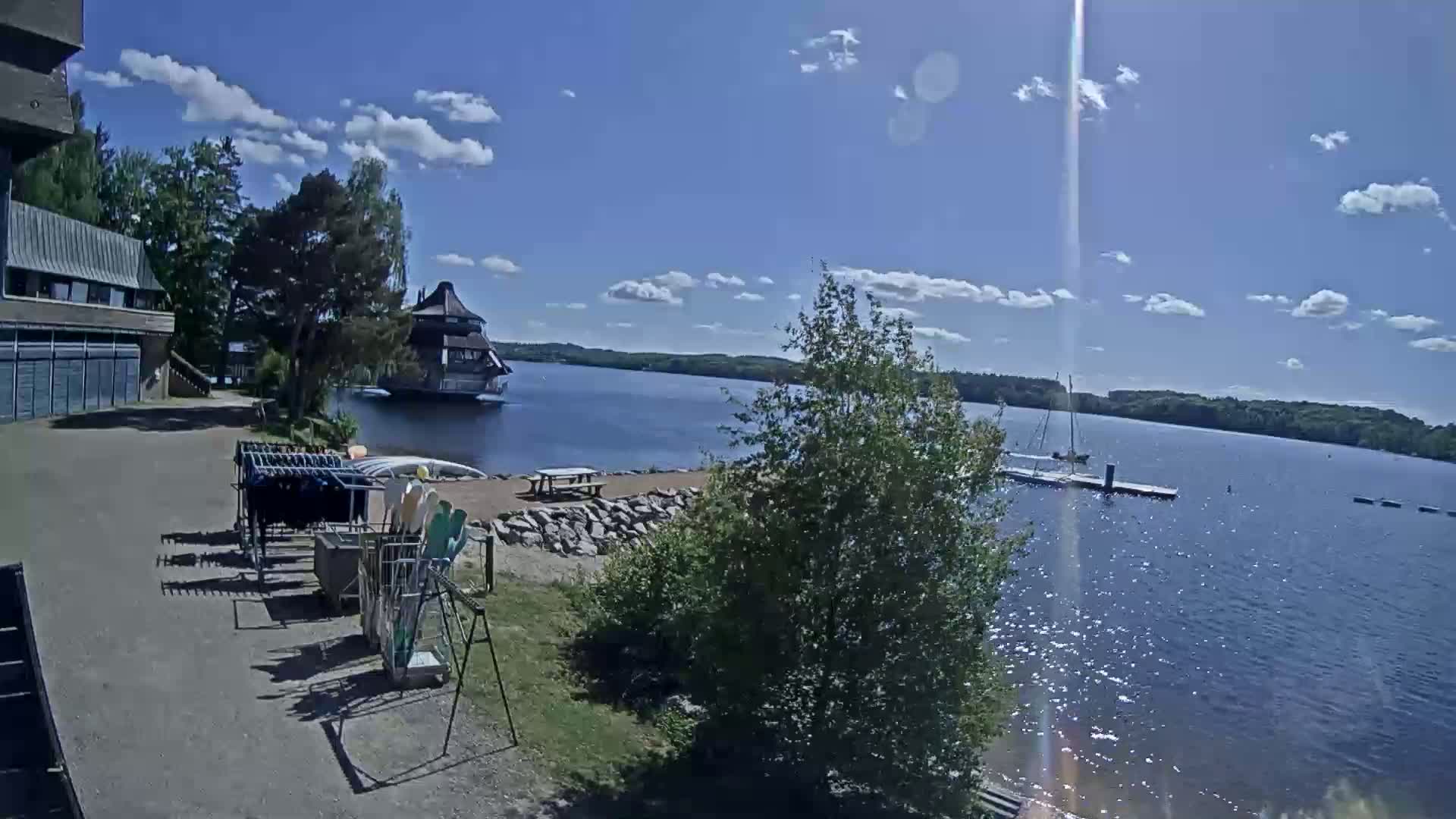 A sunny day reveals a calm lake with a building on the water, boats docked, and water sports equipment stored near a building on the shore.
