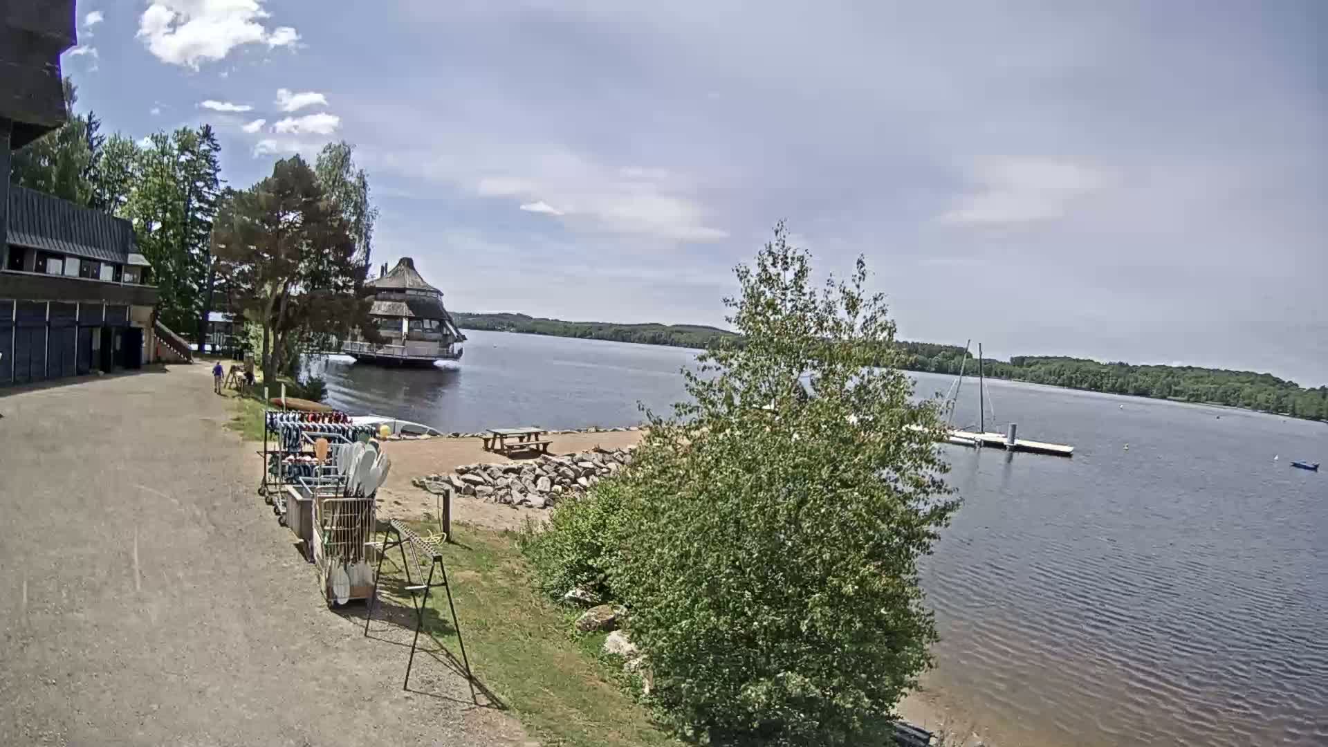 A lakeside scene on a partly sunny day shows a building, a small beach area with picnic tables, a dock with a sailboat, and various water sports equipment.