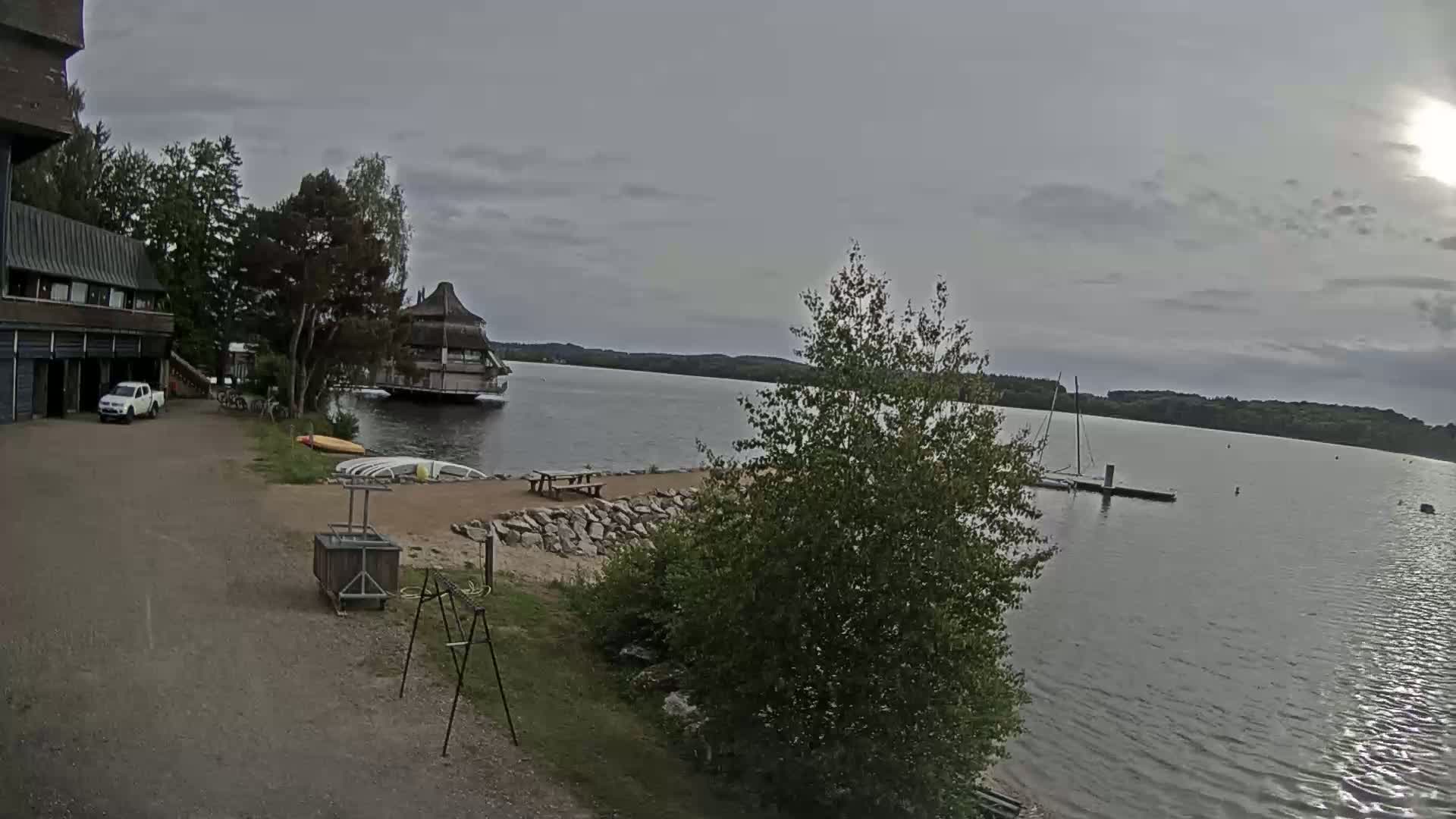 A mostly cloudy day reveals a lakeside scene with a building, a small sandy beach area, a dock with a sailboat, and a pickup truck parked nearby.
