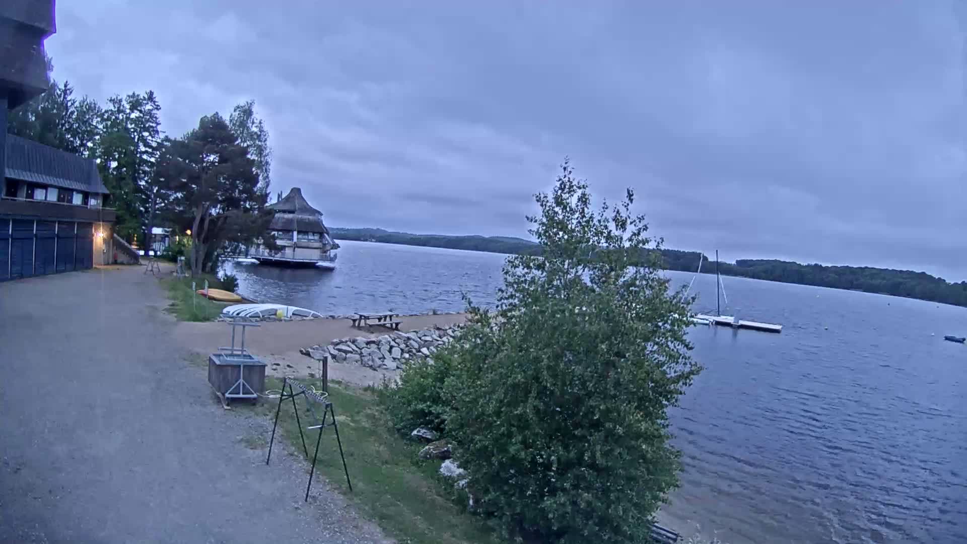 A calm lake is seen from a lakeside gravel path, with a building, a small beach area, several boats, and trees under an overcast sky.