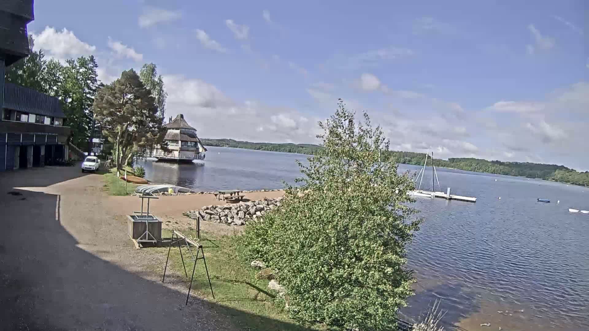 A lakeside scene on a partly cloudy day shows a building, a small beach area with picnic tables, a boat dock with sailboats, and a large tree in the foreground.