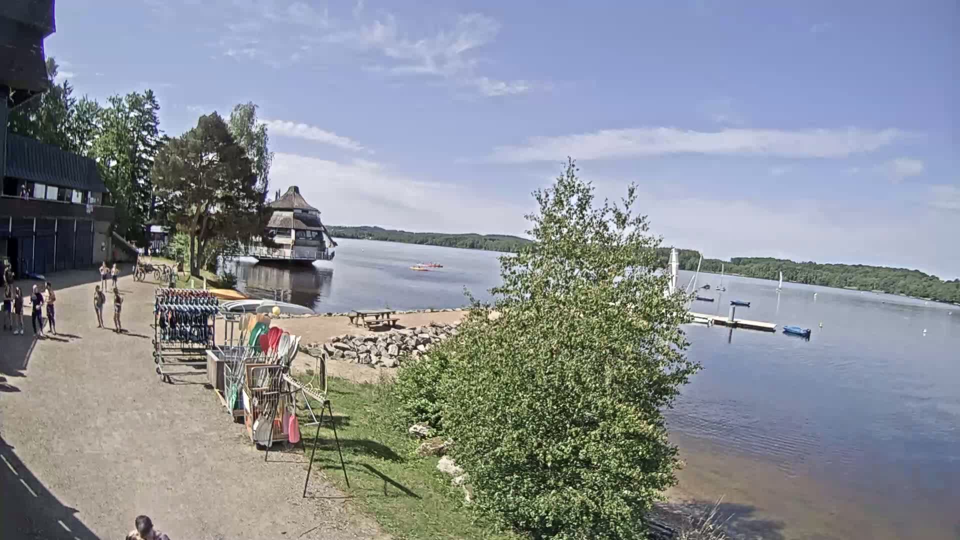 A sunny day at a lakeside recreational area shows people near a building with watercraft rentals, a small beach area with picnic tables, and sailboats on the calm water.