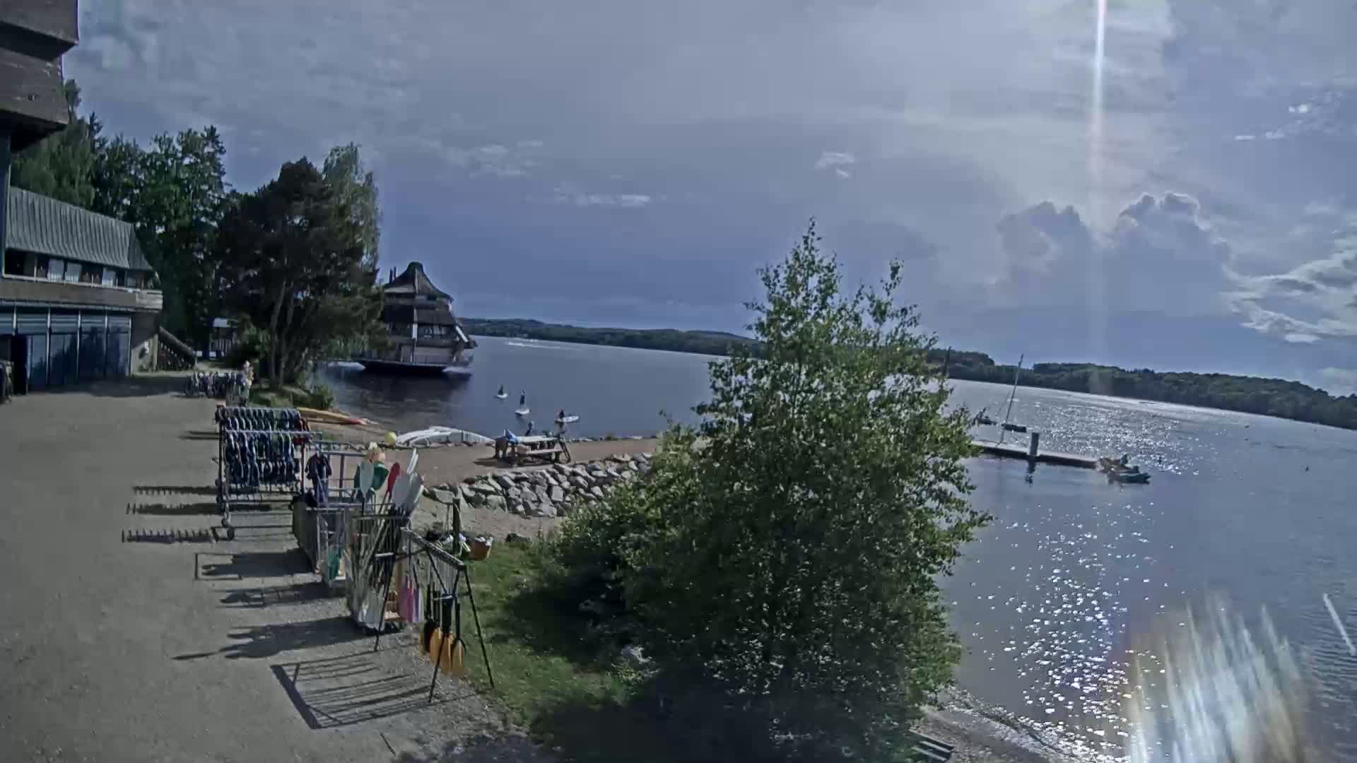 A lakeside scene on a partly sunny day shows a building, people engaging in watersports, and various watercraft, with paddleboards and other equipment stored nearby.