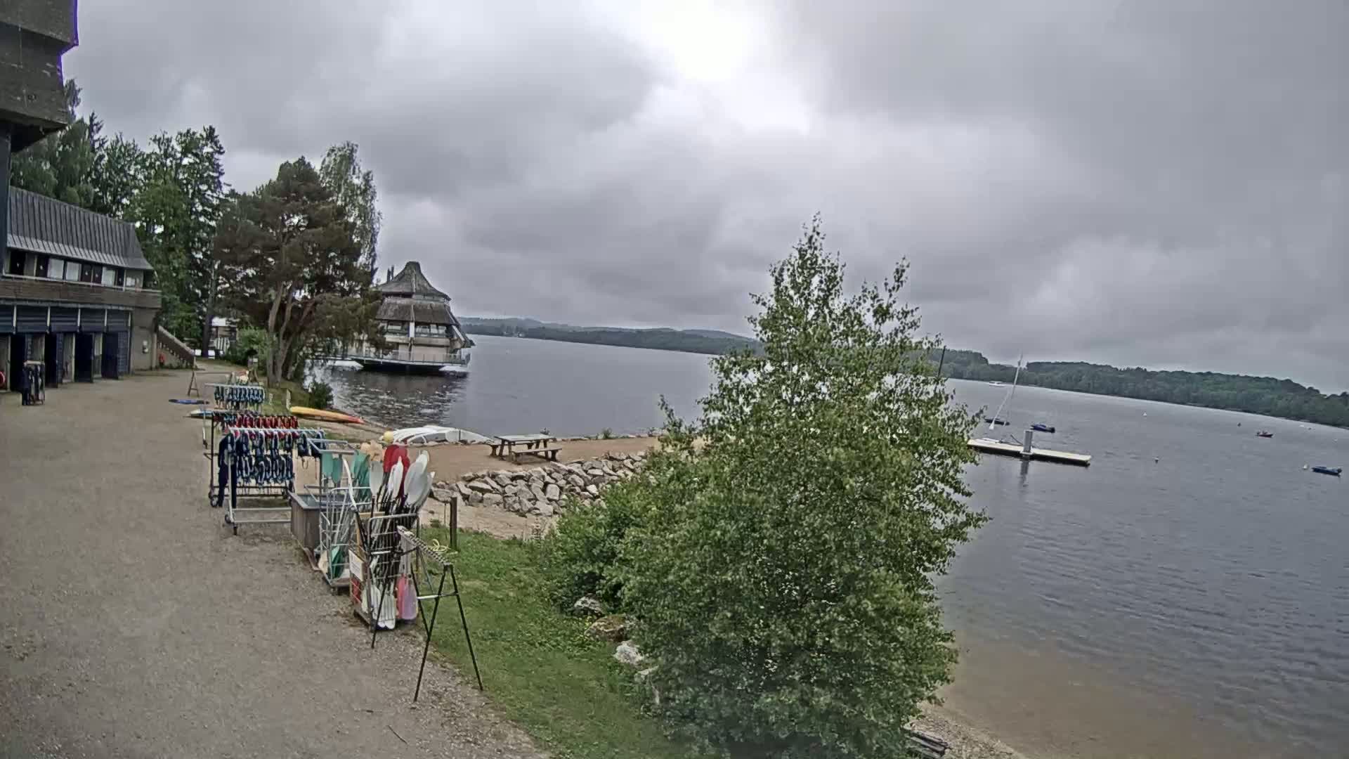 Kayaks and other watercraft are stored near a building overlooking a calm lake under an overcast sky.