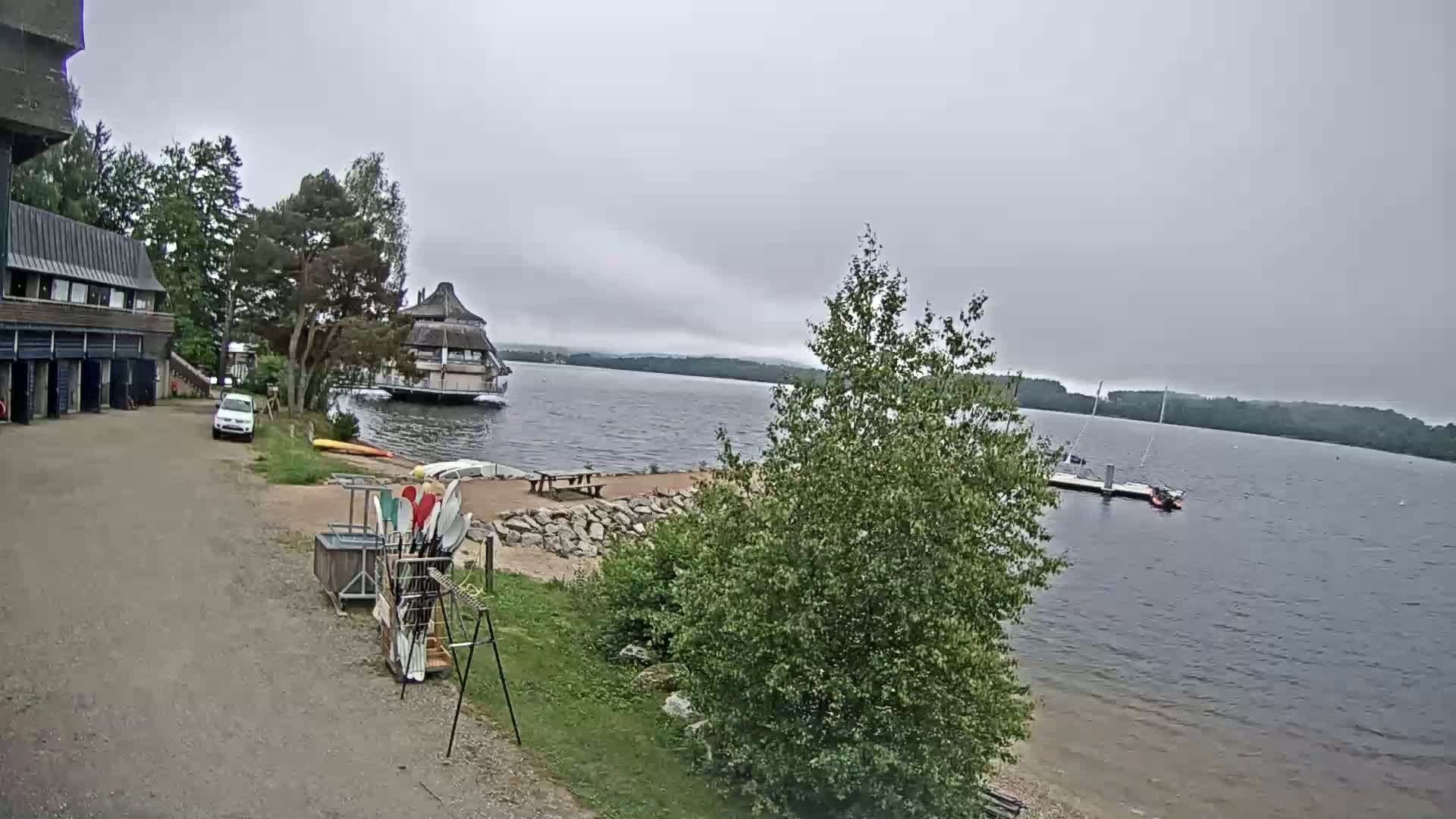A lakeside view on an overcast day shows a building with kayaks and a small car in a parking area, a restaurant on the water, and sailboats moored on a dock.