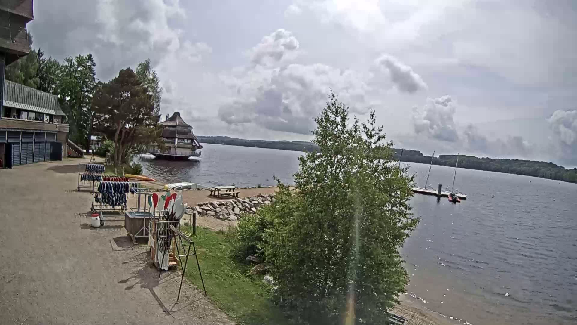 A lakeside scene under a partly cloudy sky shows a building, watercraft, and equipment for watersports near a sandy shore.