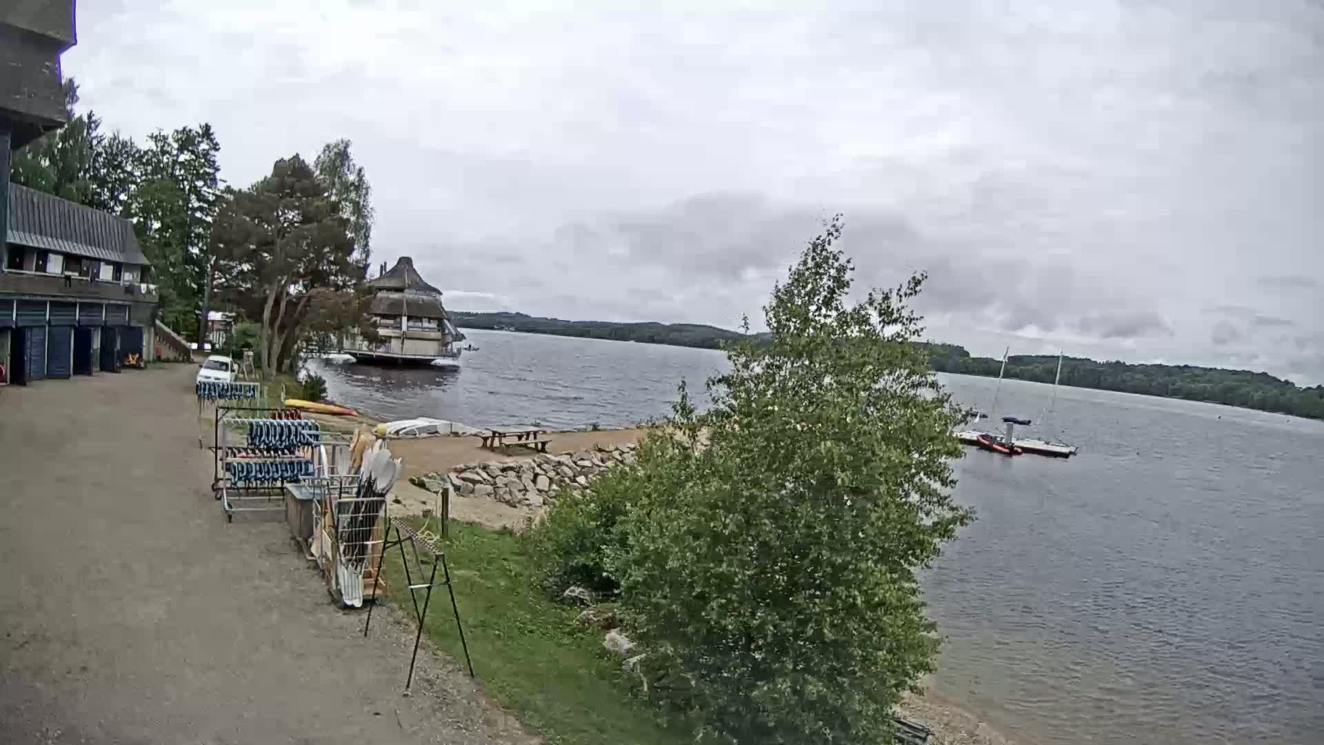 A lakeside scene on an overcast day shows a building with a small parking area, watercraft storage racks, and a calm lake with boats in the distance.