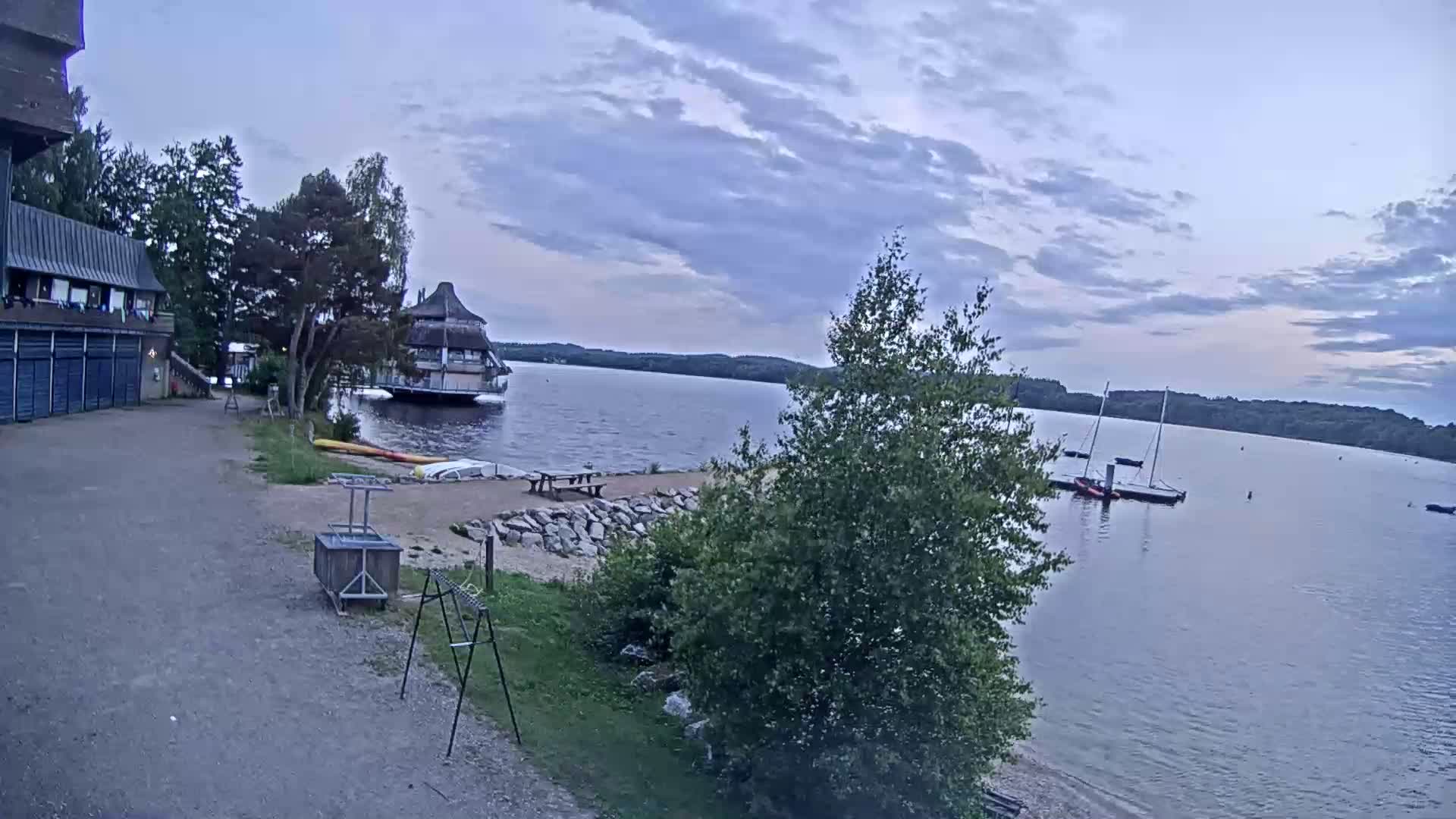 A lakeside scene under a cloudy sky shows a building, a small beach area with picnic tables, kayaks, sailboats on the water, and green vegetation.