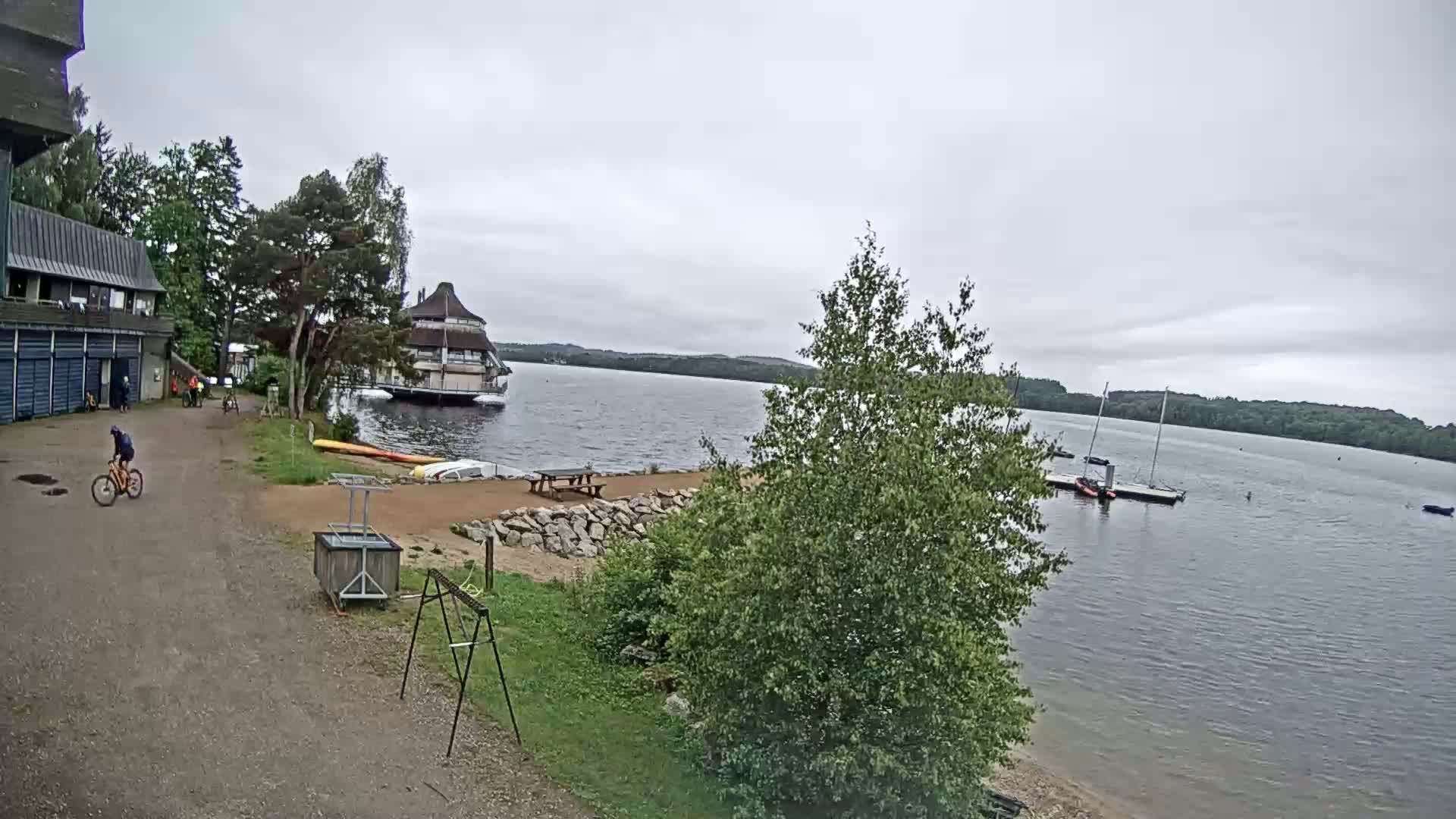 A person bikes along a path near a lake under an overcast sky, past a building and a sandy area with boats and picnic tables.