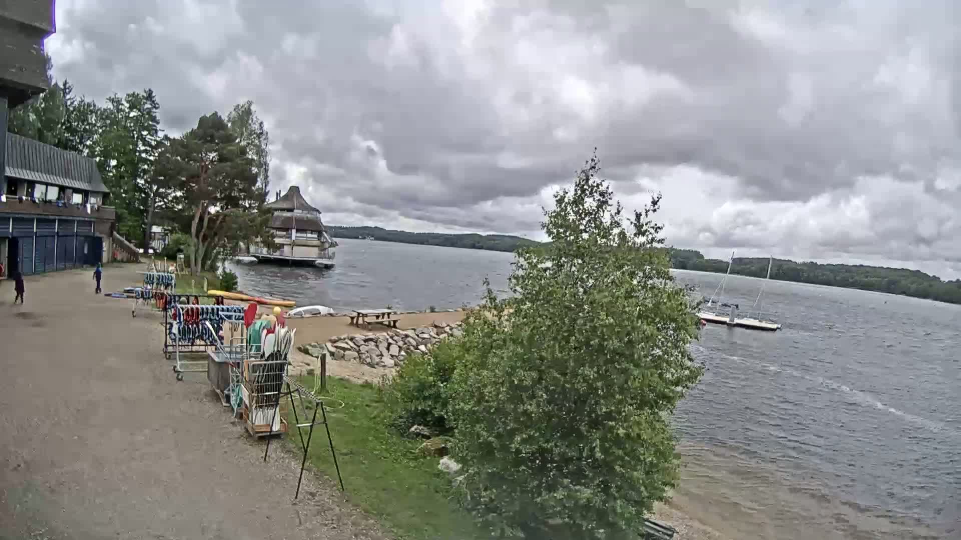 A lakeside scene under a cloudy sky shows a building with kayaks and paddleboards stored outside, a small sandy beach area, and boats on the water.