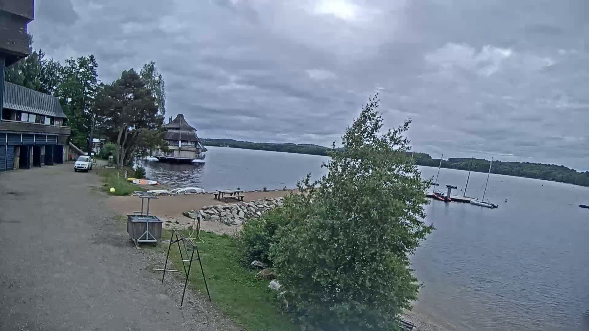 A lakeside view on an overcast day shows a building with garages, a small white car, several sailboats moored on the water, a circular building on the lake, and a sandy area with picnic tables.
