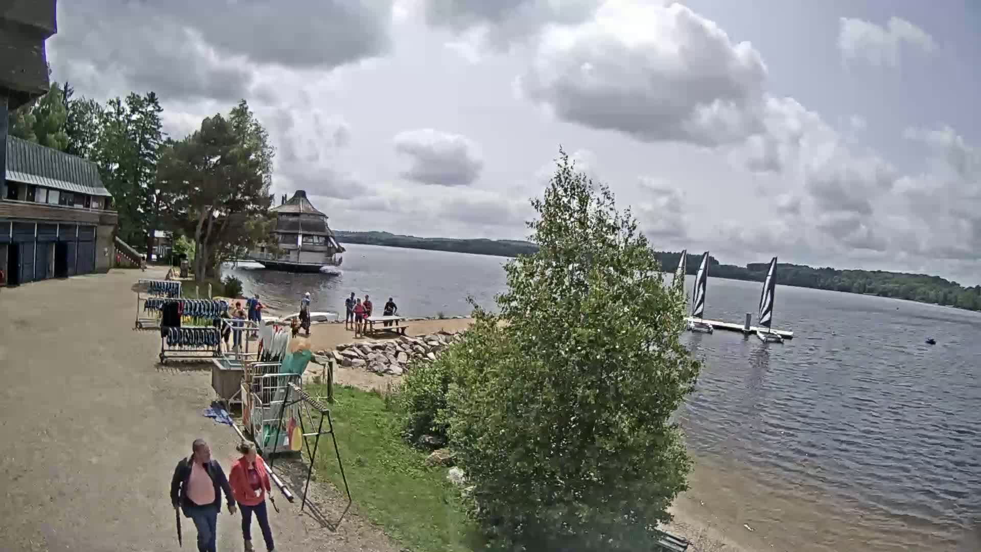 A partly cloudy day at a lakeside recreational area shows people gathered near a building and on the sandy shore, with sailboats on the water.