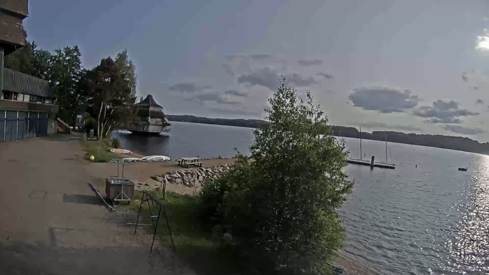 A lakeside scene on a partly cloudy day shows a building, a floating structure, a small sandy beach area, some trees, and boats on a calm lake.