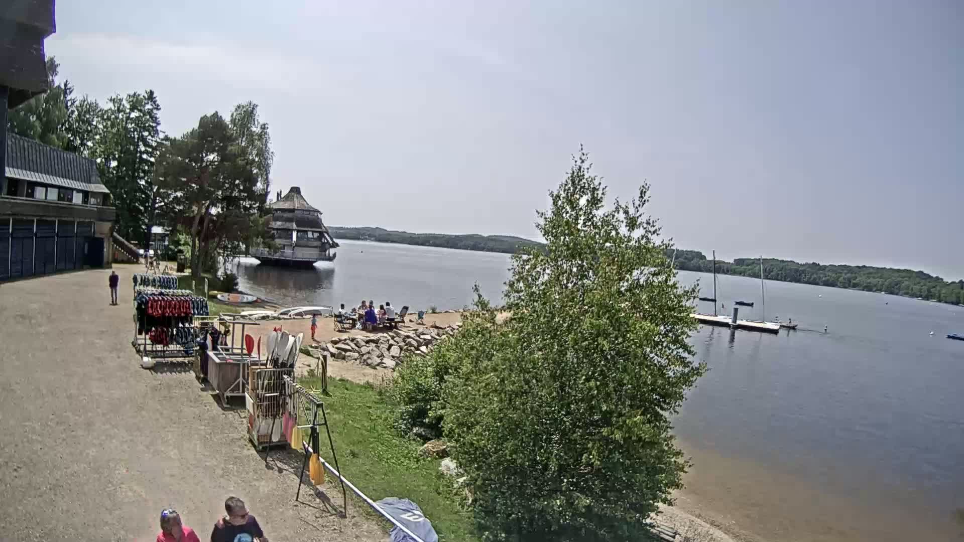 A sunny day at a lakeside recreational area shows a sandy beach with people sitting at picnic tables, boats on the water, and equipment rentals near a building.