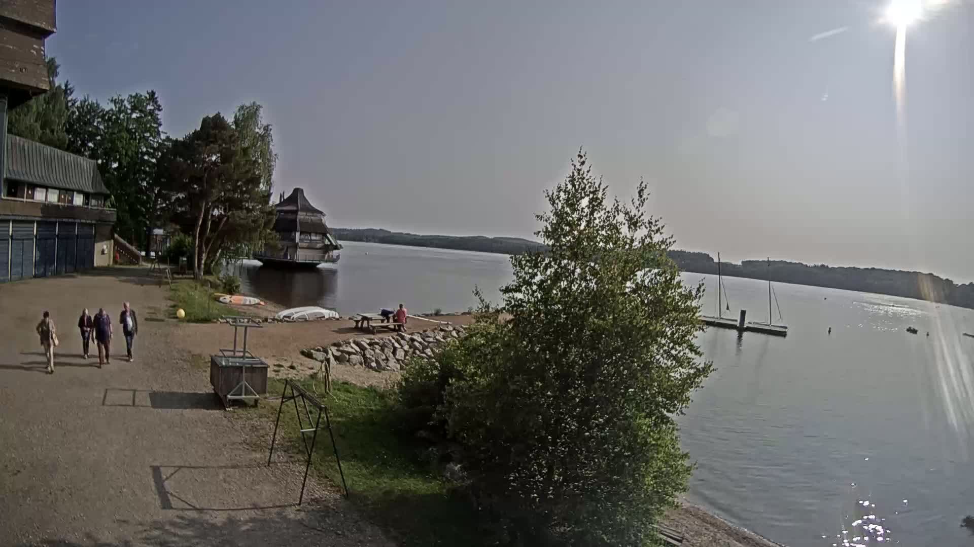 A sunny day at a lake shows a group of people walking near a building and a floating structure, with boats and a person sitting at a picnic table on the shore.