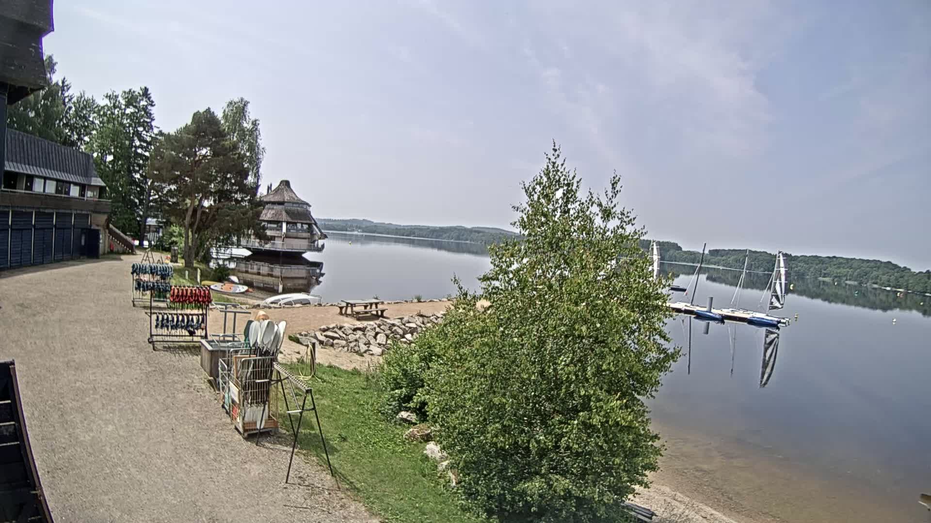 A calm lake is seen from a lakeside building with several boats docked and kayaks stored near a sandy shore under a mostly clear sky.