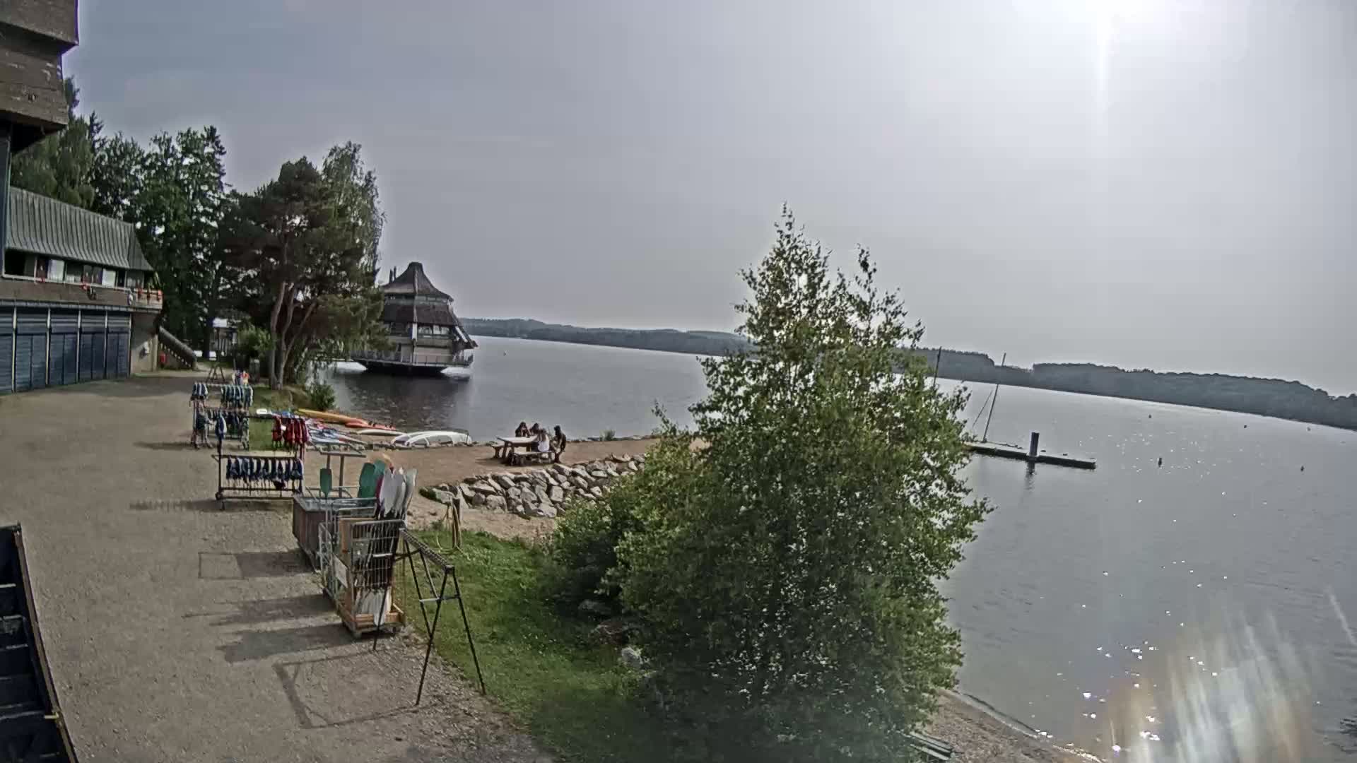 A lakeside scene on a sunny day shows a building, a small beach with people sitting at a picnic table, and various watercraft and equipment near a parking area.