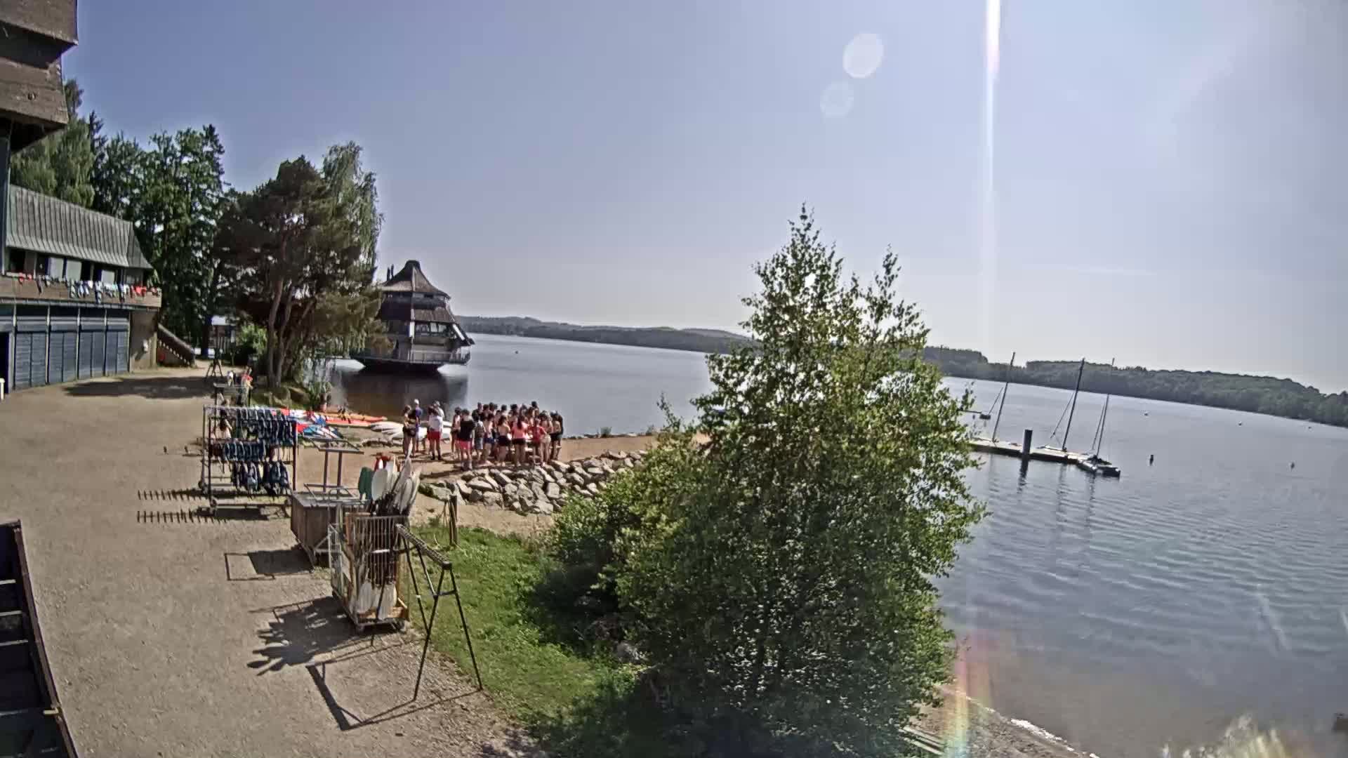 A sunny day at a lakeside recreation area shows a group of people gathered near a building, various watercraft, and equipment racks.