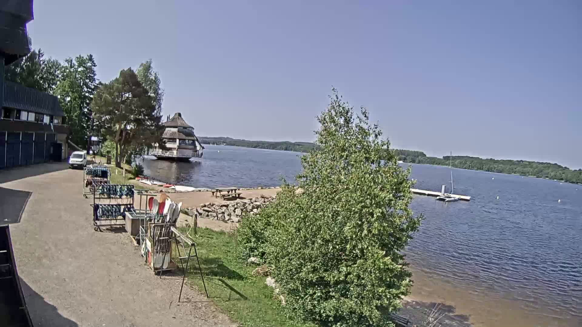 A lakeside view on a sunny day shows a building, a small beach area with picnic tables, and watercraft, with equipment racks and a vehicle parked nearby.