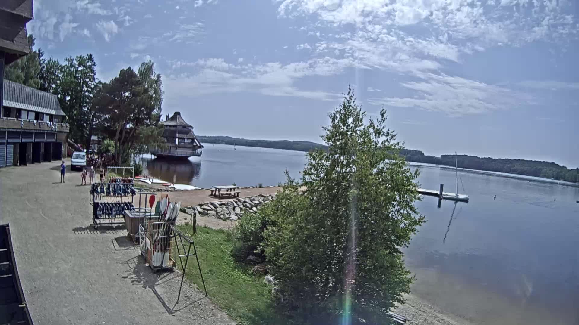 A calm lake under a partly cloudy sky is seen from a lakeside building's patio area, where people are gathered near racks of water sports equipment.