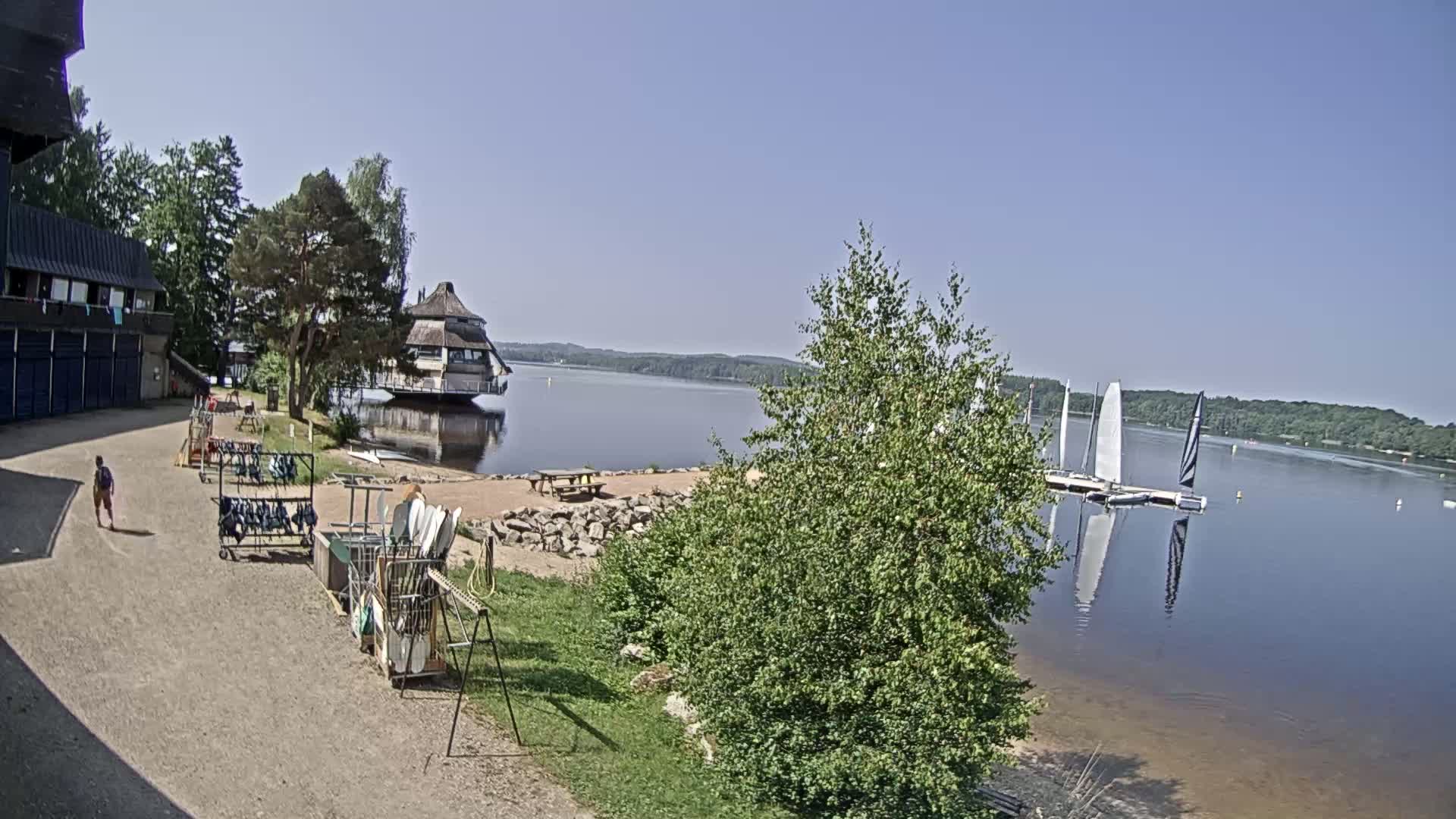A person walks past a lakeside building and equipment storage area on a sunny day, with sailboats visible on the calm water.