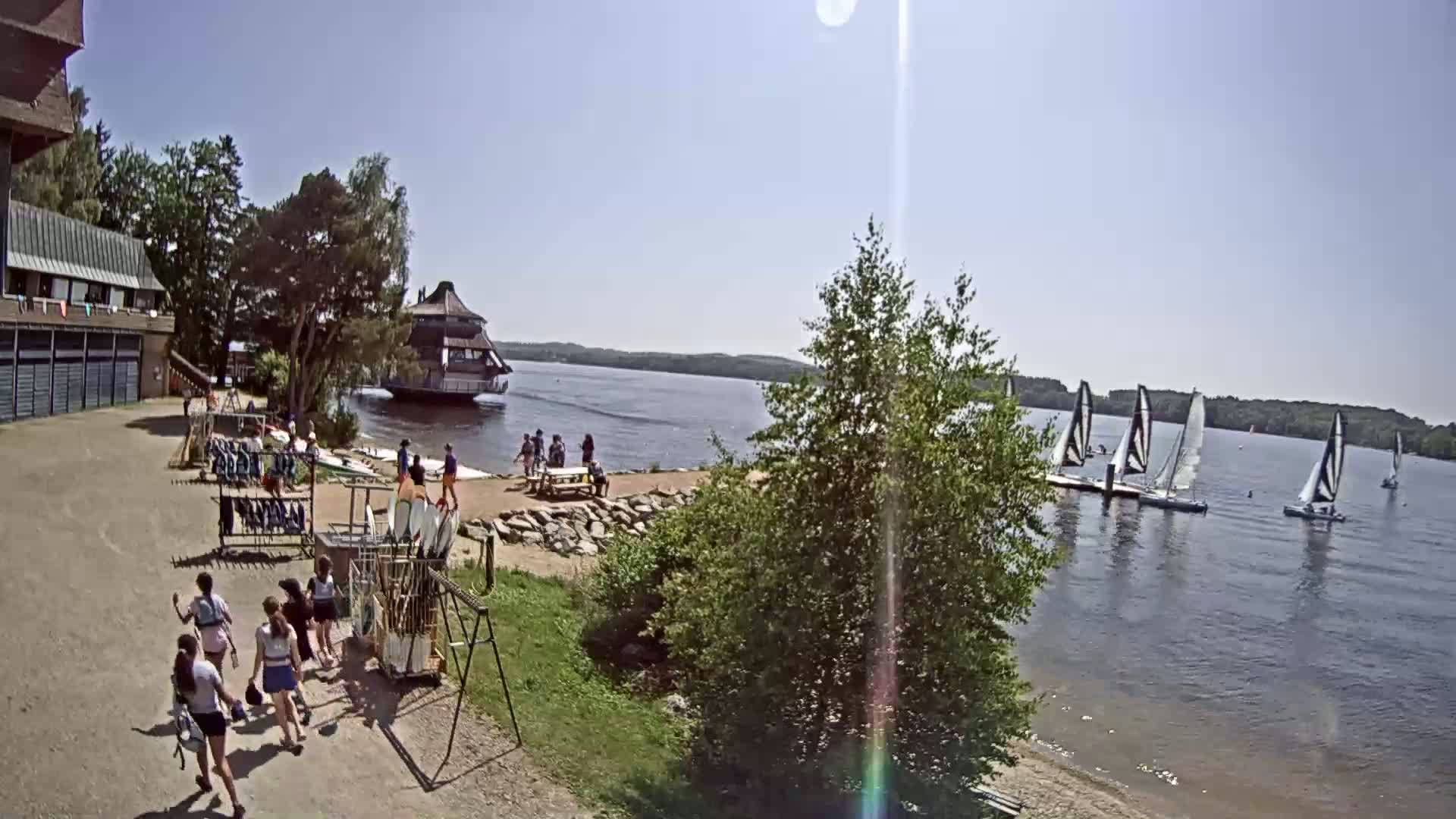 On a sunny day, several people walk near a lakeside building and equipment storage area, while sailboats are visible on a calm lake in the background.