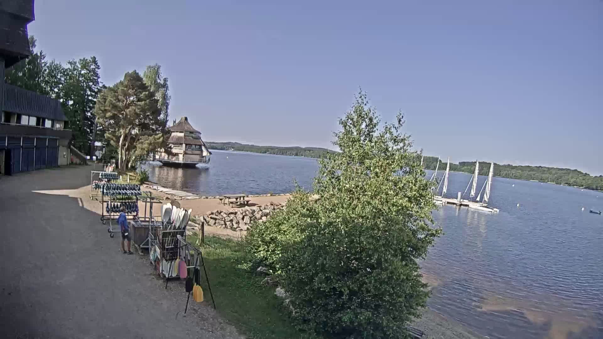 A sunny day at a lakeside facility shows a building with watercraft and equipment, along with several sailboats moored on the calm water.