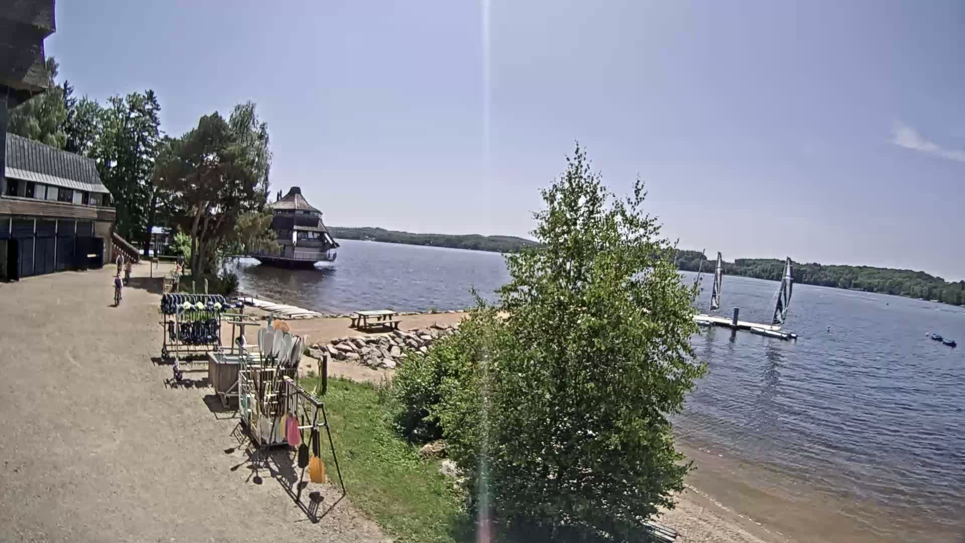 A lakeside scene on a sunny day shows a building, a dock with sailboats, and various water sports equipment near a sandy shore with green vegetation.