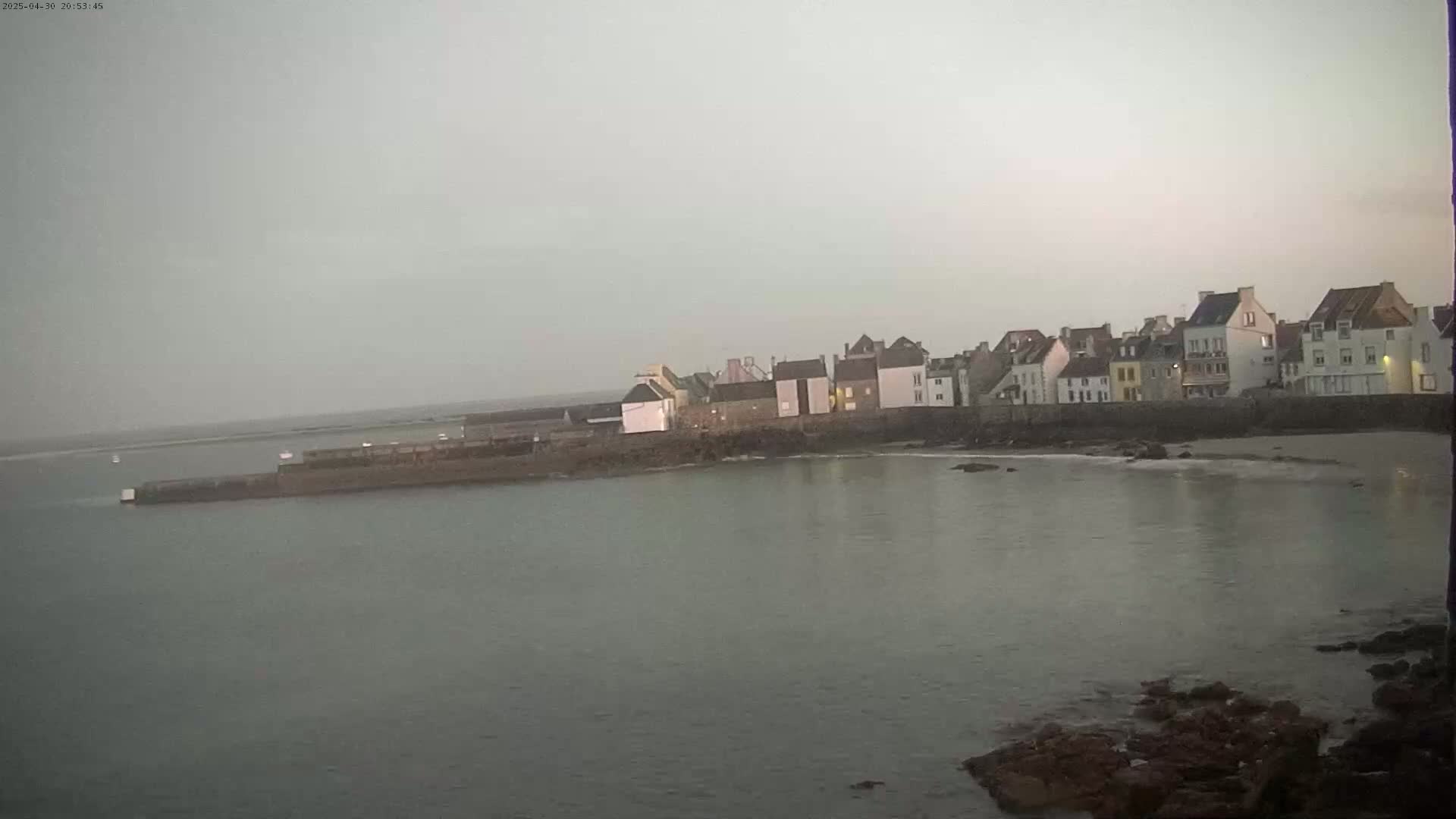 A row of buildings sits on a rocky coastline next to calm, gray water under an overcast sky.