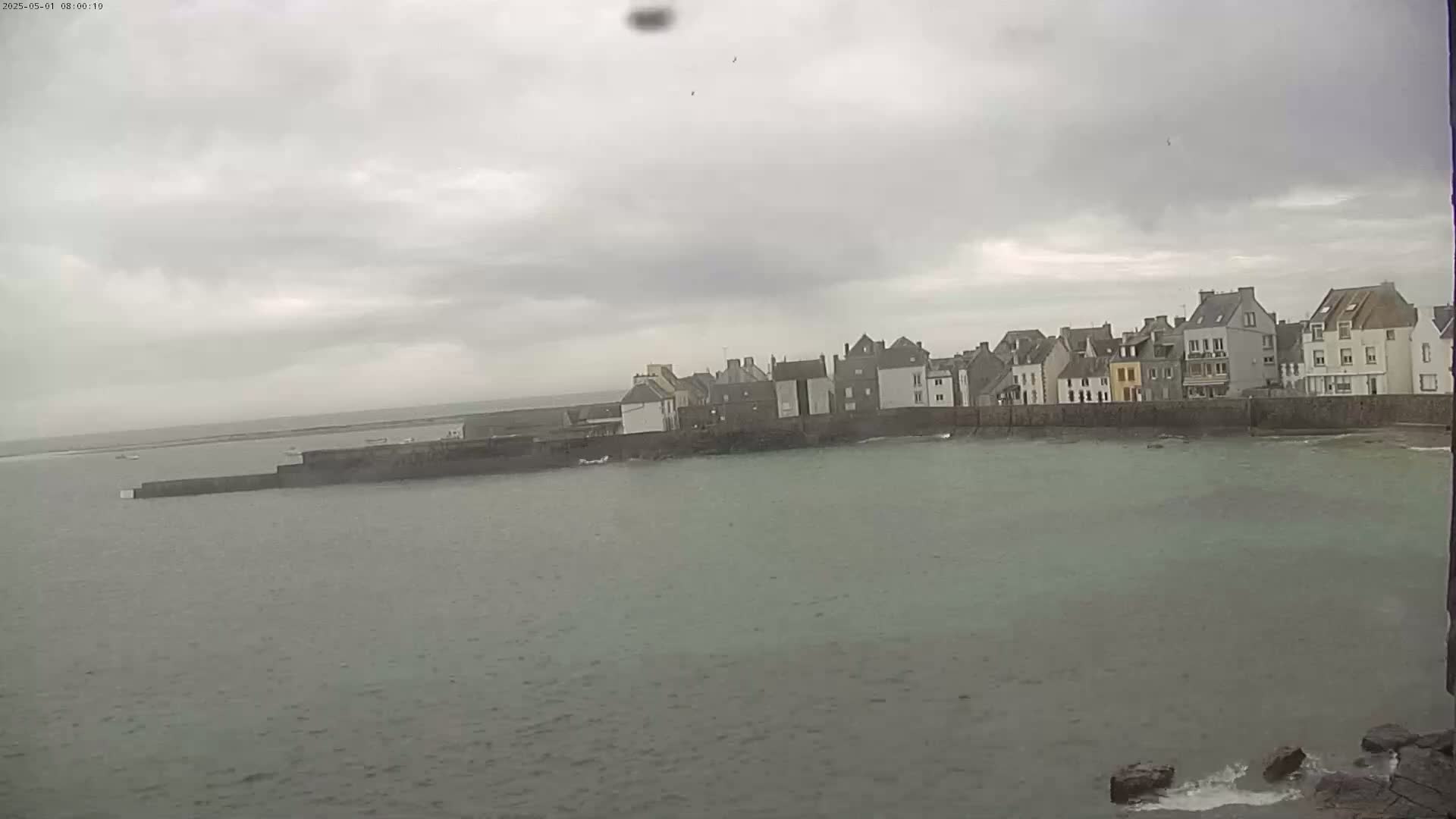 A row of buildings sits behind a seawall overlooking a calm, gray body of water under an overcast sky.