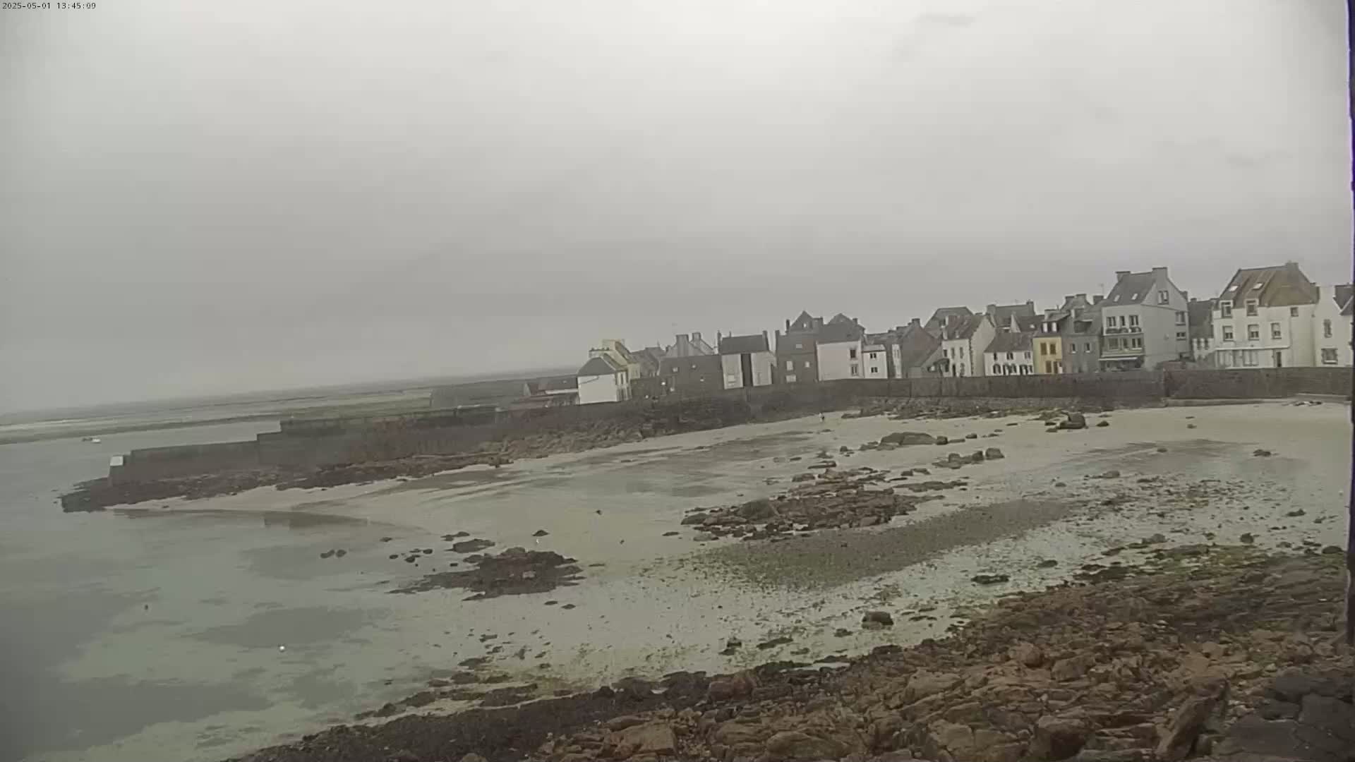 A row of buildings sits behind a sandy beach and rocky shoreline on an overcast day.