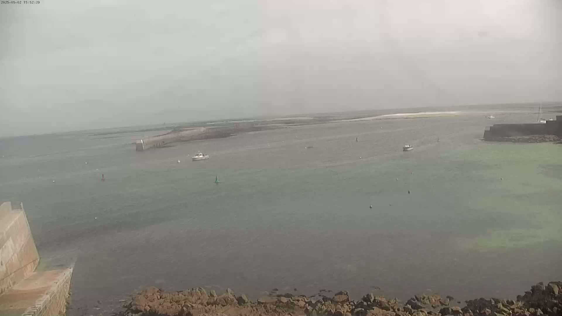 A hazy, overcast day reveals a calm body of water with several small boats, rocky shores, and a stone structure extending into the sea.