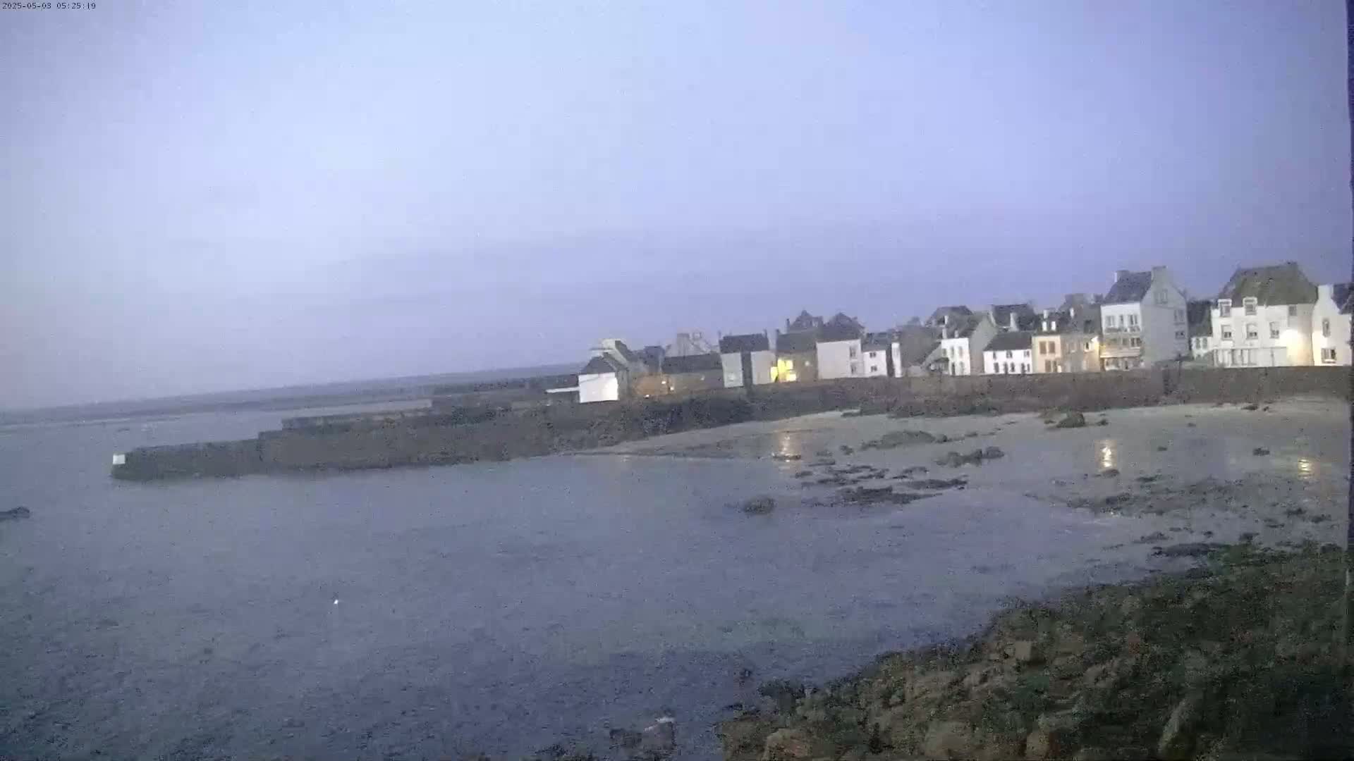 A row of buildings sits on a rocky shoreline next to calm, shallow water under a dusky sky.