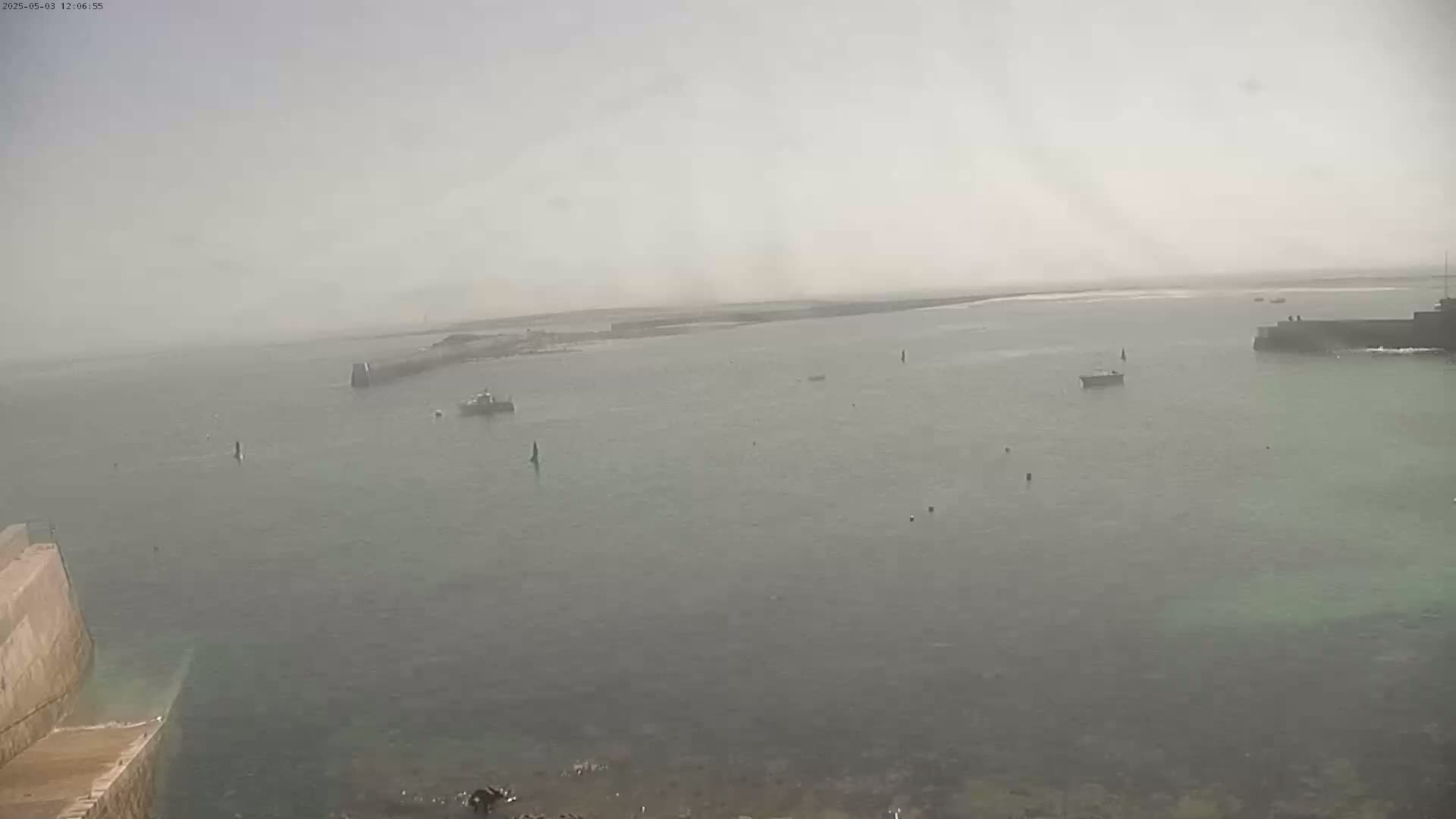 A hazy, overcast day reveals a calm body of water with several small boats and harbor structures.