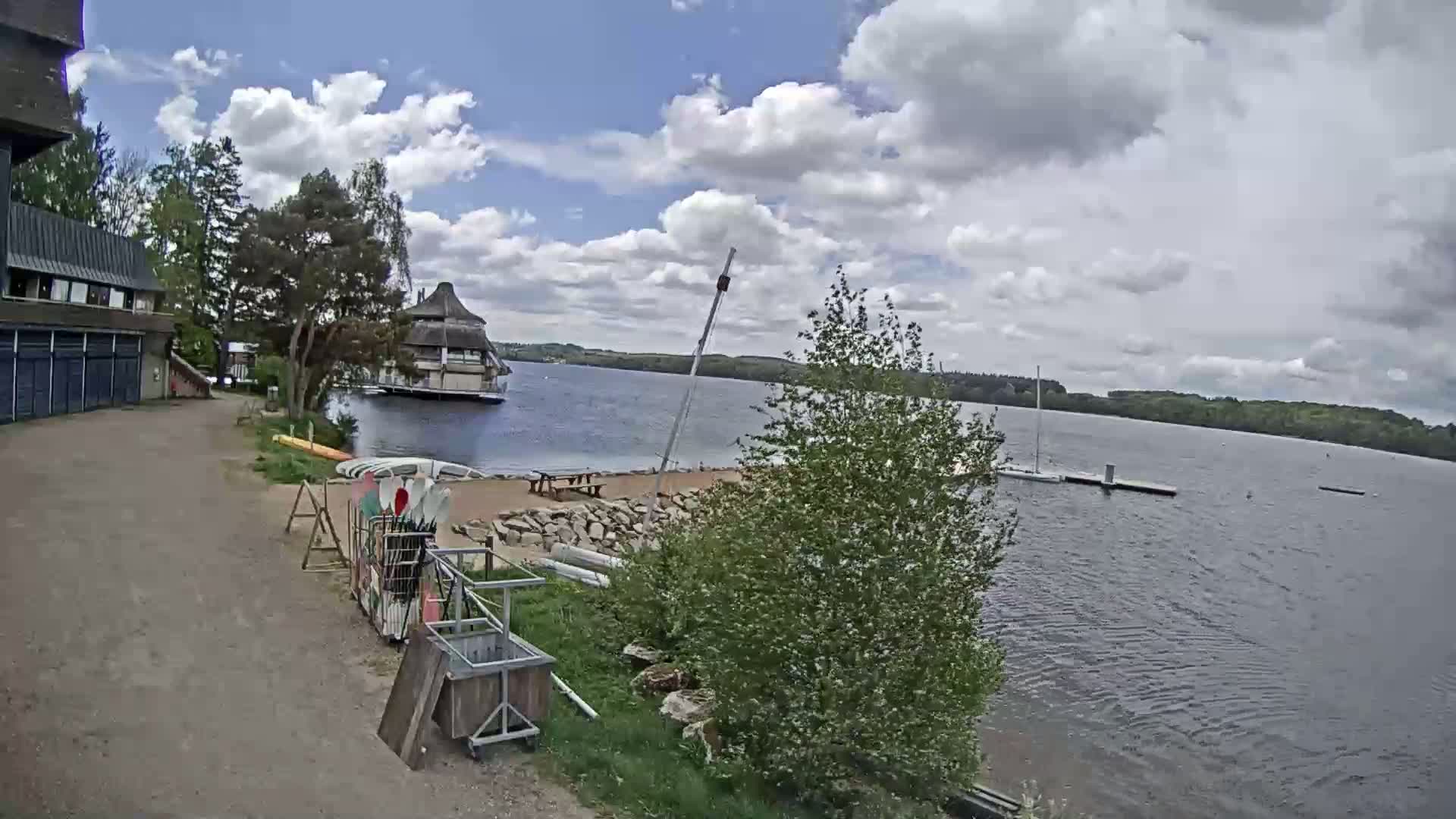 A partly cloudy day shows a lakeside view with a building, boats, and kayaks near a sandy beach area.
