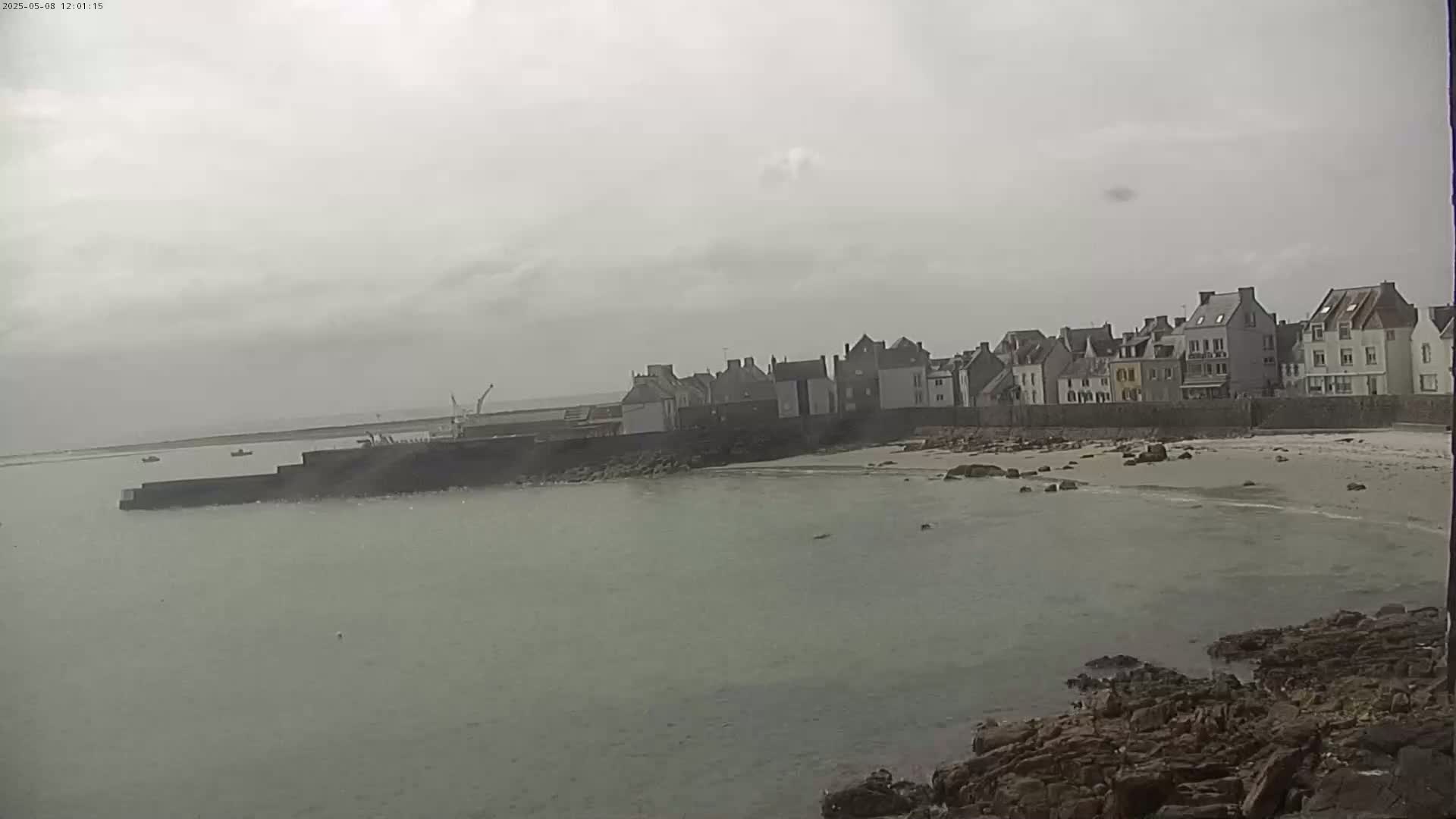 A coastal town with multi-story buildings is seen on an overcast day, with calm water in the foreground and a small sandy beach.
