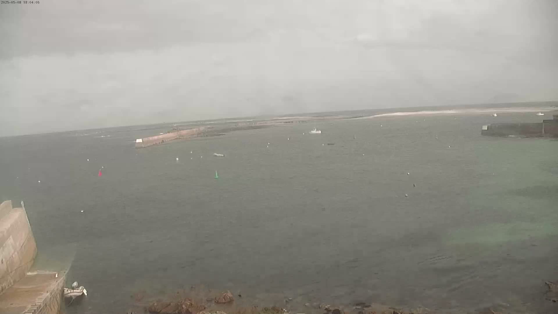 A calm, overcast day shows a harbor with several small boats, a stone wall, and a stretch of land in the distance.