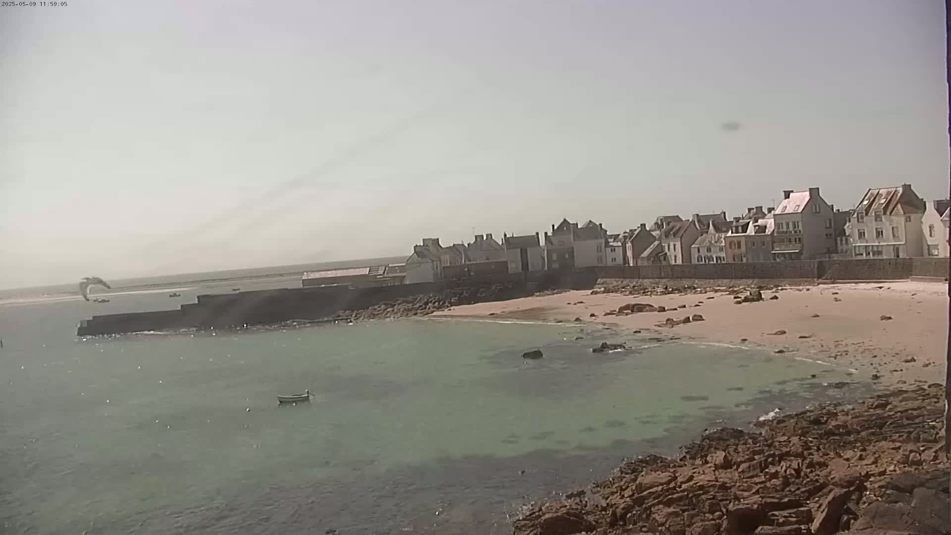 A sandy beach with calm, shallow, green-tinged water borders a row of buildings under a hazy sky.
