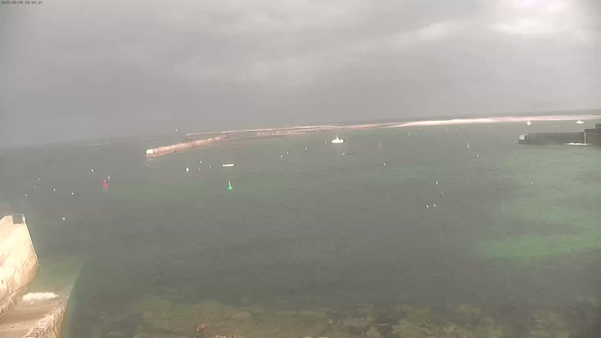 A hazy, overcast day reveals a calm body of water with several small boats and buoys, partially enclosed by low stone walls.
