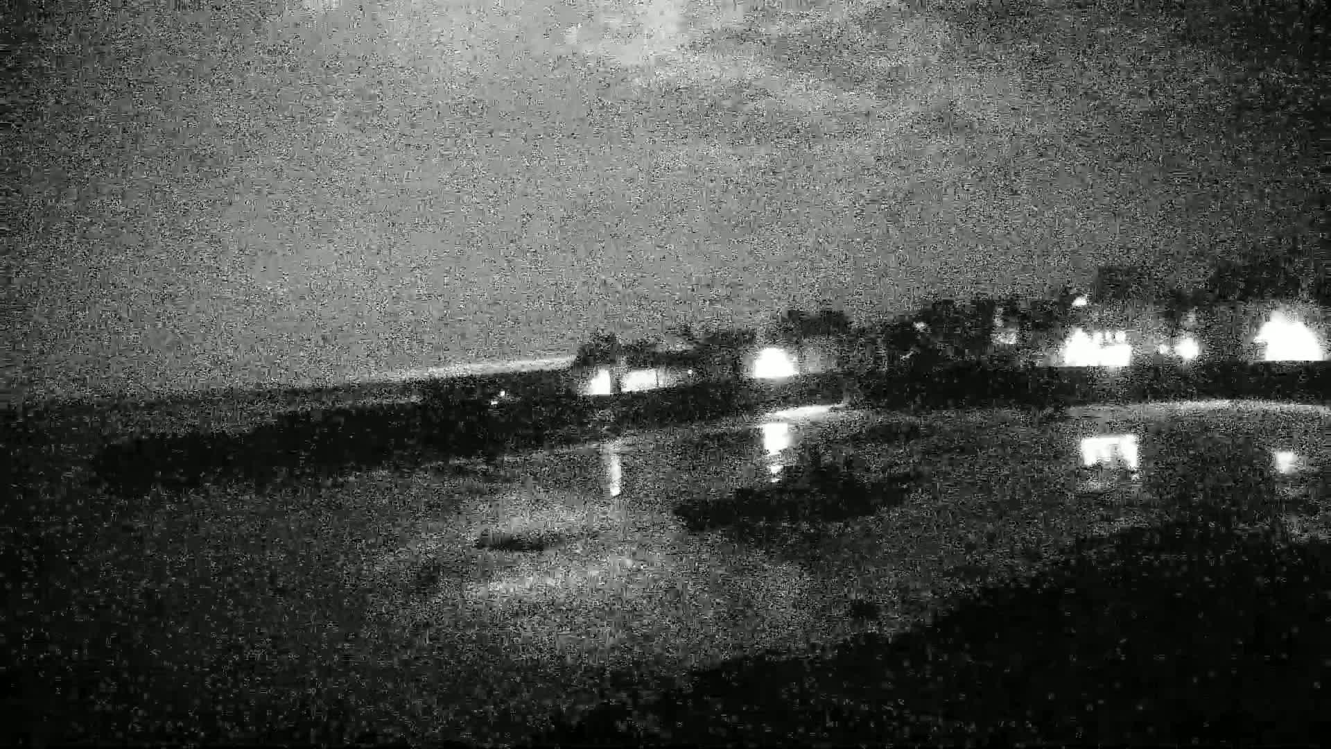 A nighttime, low-light view shows a line of buildings with lights on near a dark body of water under a cloudy sky.
