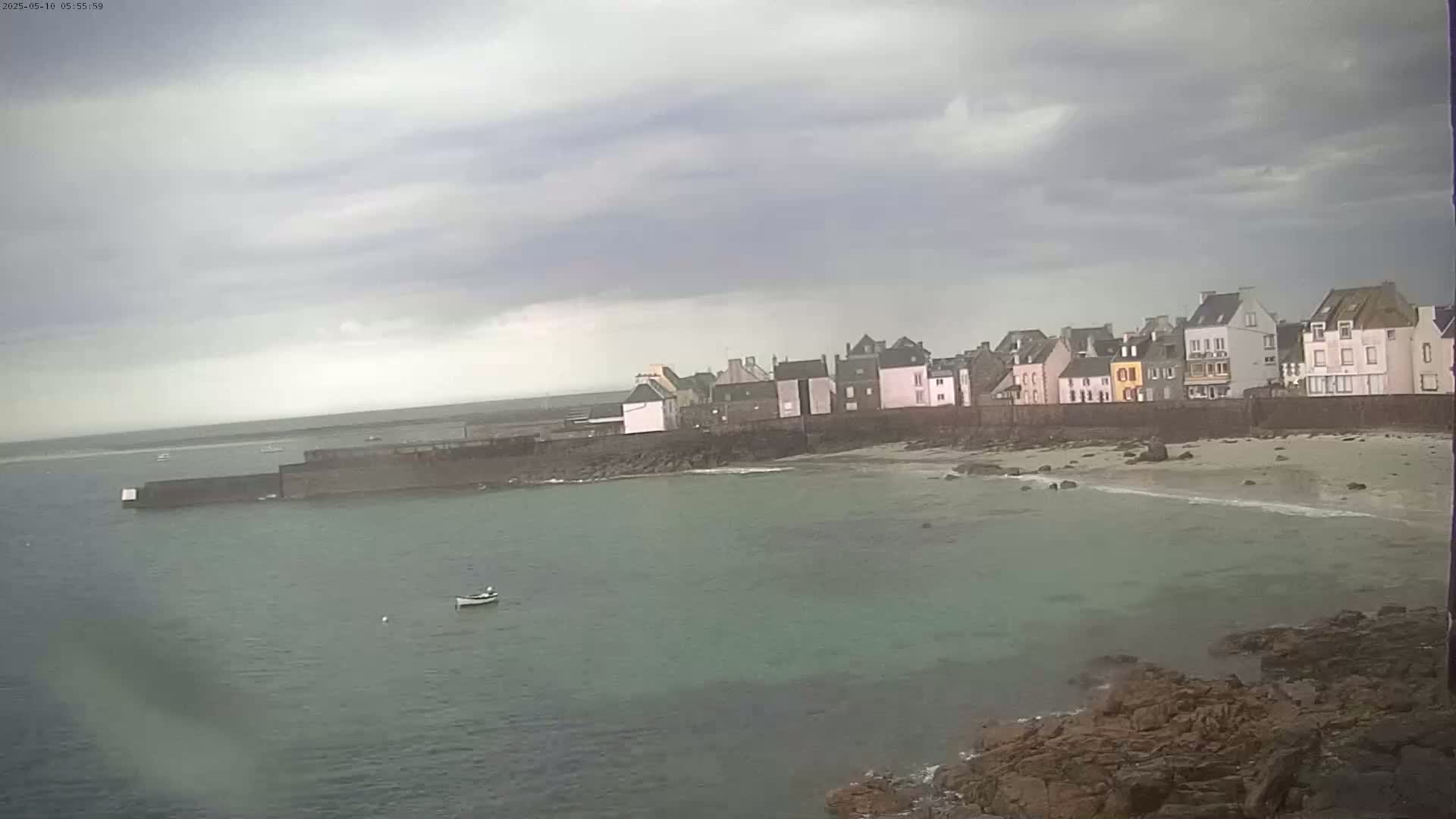 A small, coastal town with overcast skies sits beside a calm body of water, a single small boat visible in the bay.