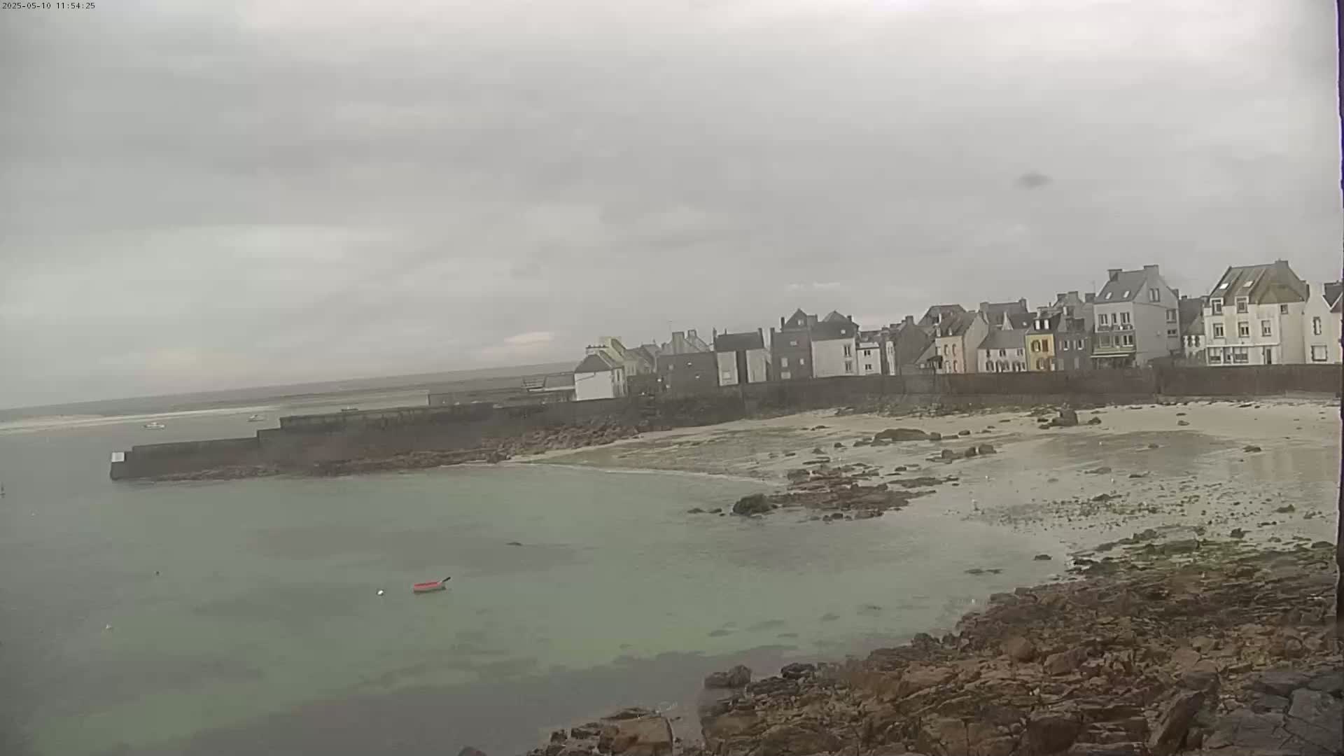 A cloudy day reveals a coastal town with a sandy beach and calm, shallow water, partially enclosed by a low stone wall.