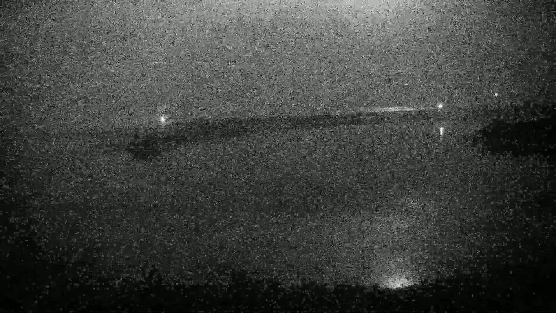 A nighttime, grainy image shows a dark body of water with faintly visible lights along a distant shoreline.