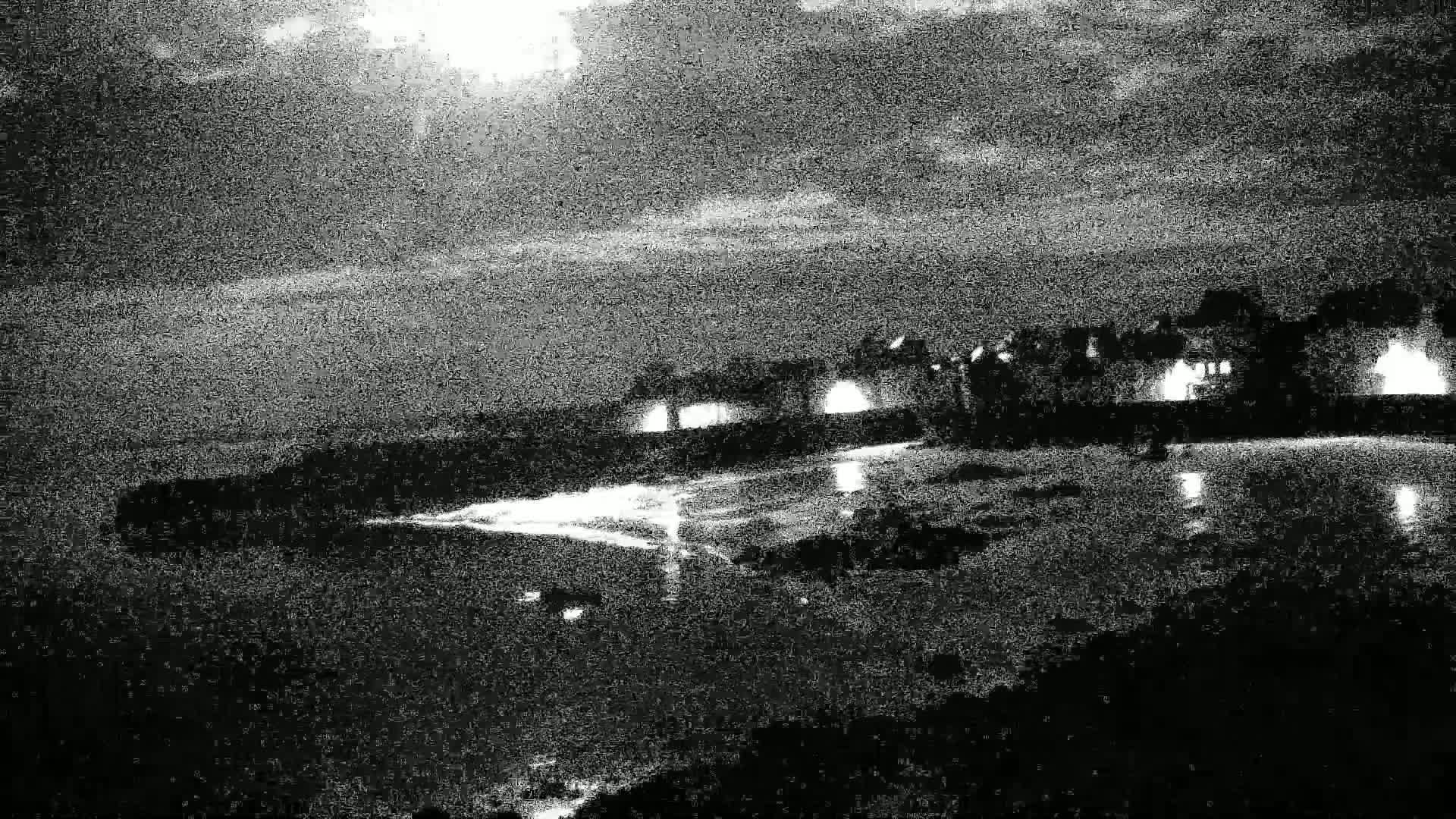 A moonlit nighttime scene shows a dark shoreline with illuminated buildings reflected in calm water under a cloudy sky.