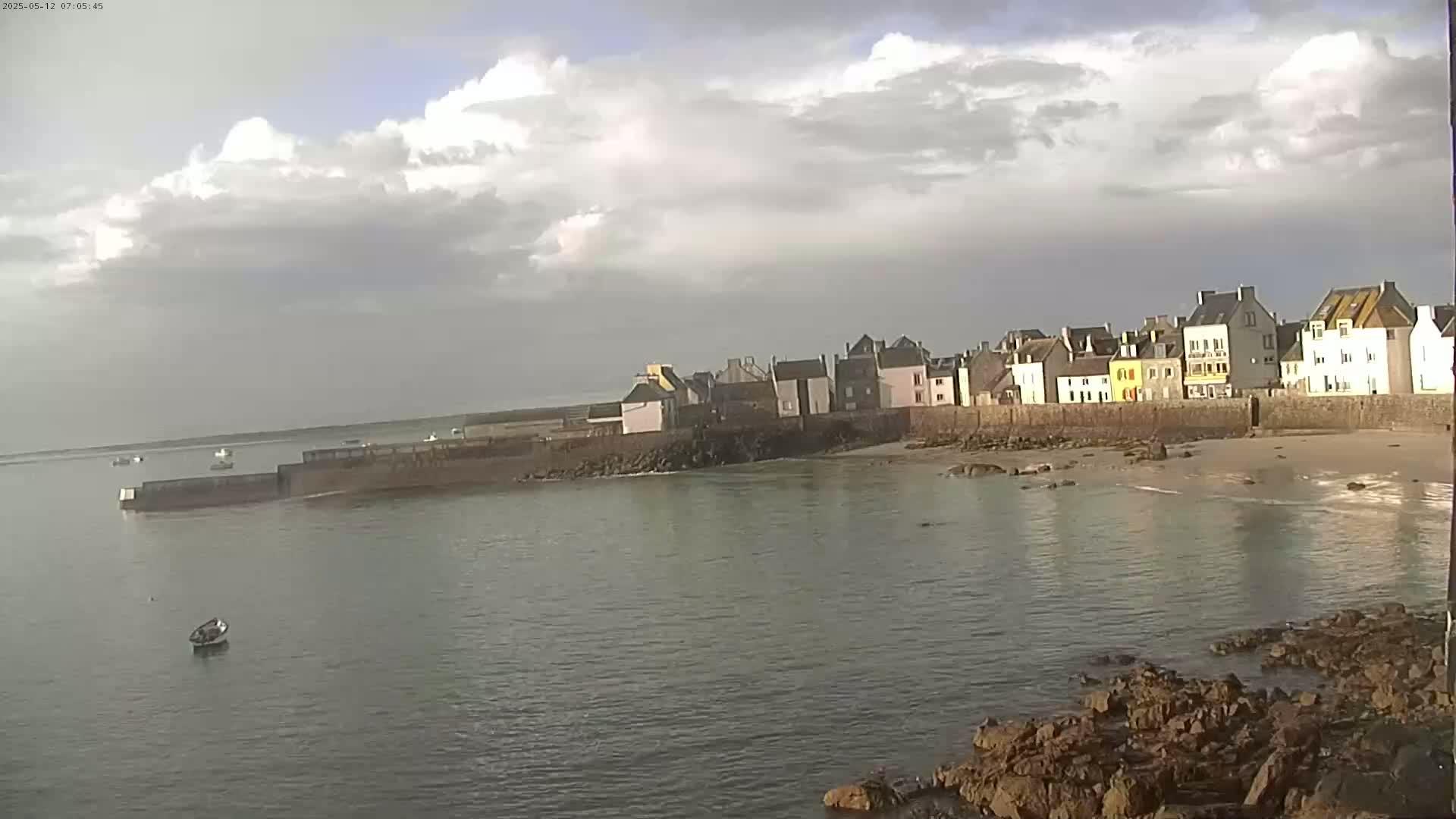 A row of buildings sits along a stone wall bordering a calm body of water under a partly cloudy sky.