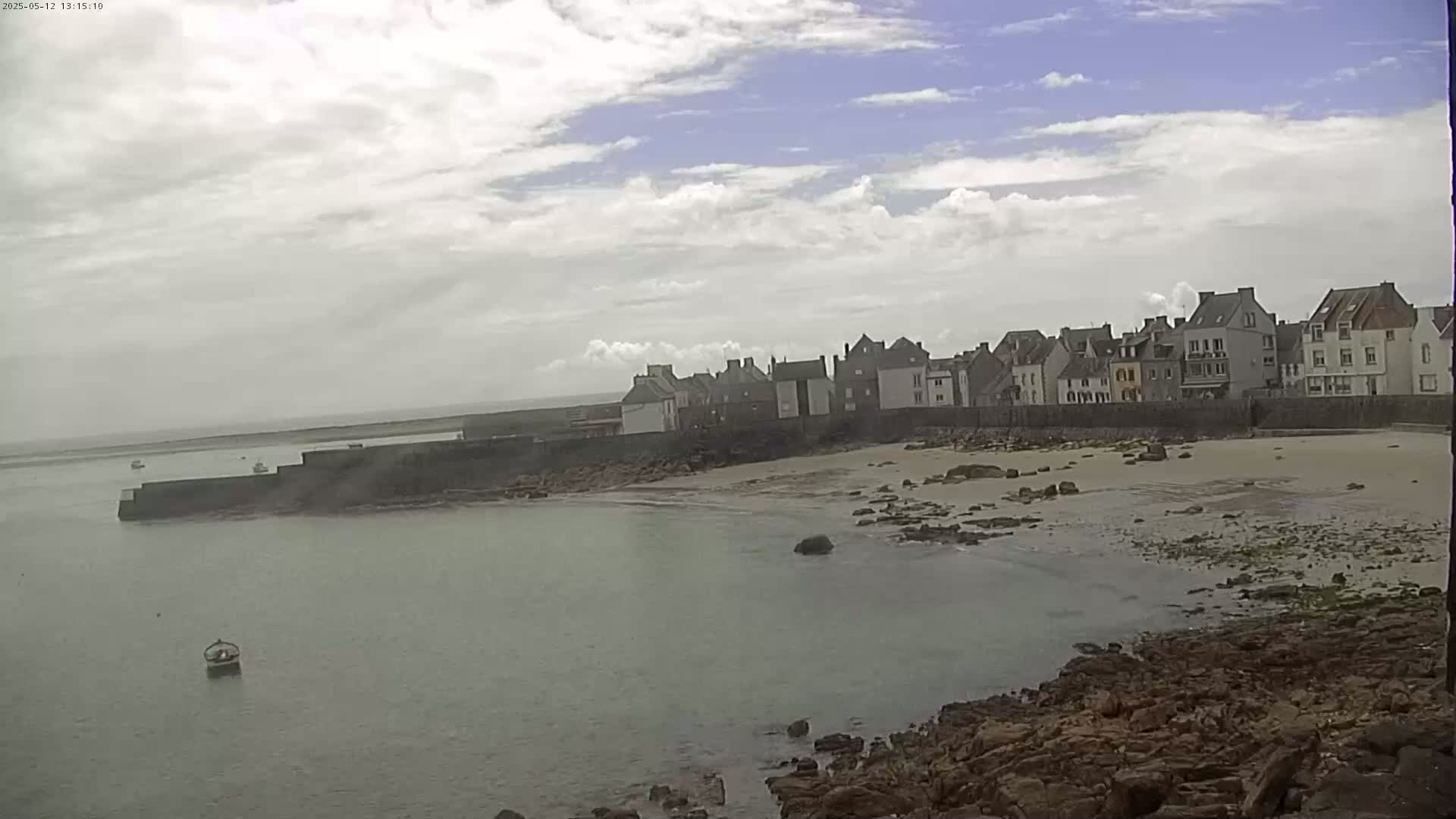 A small town with stone buildings sits along a sandy beach and calm, light-green water under a partly cloudy sky.